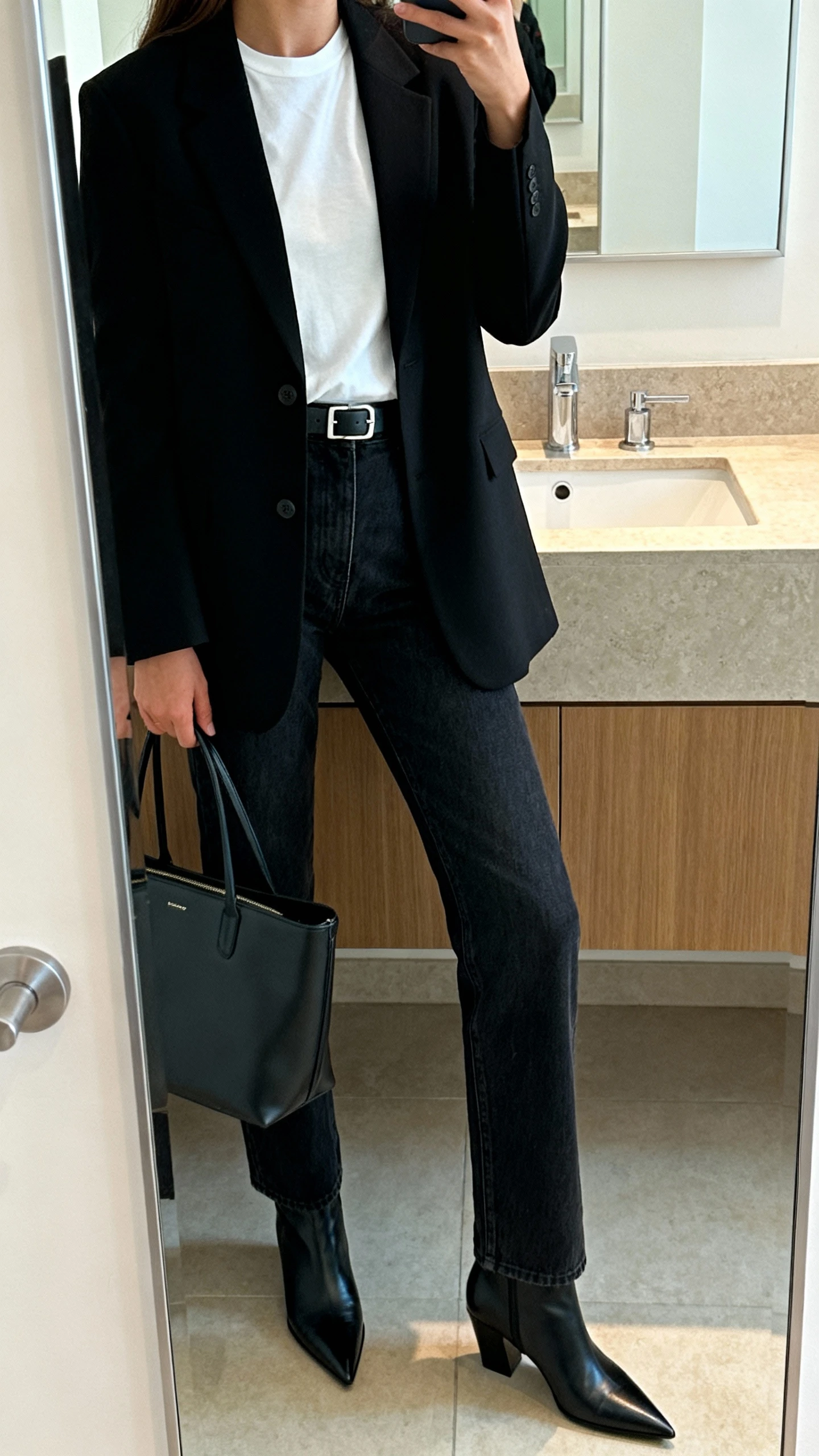 iPhone mirror selfie of a woman in elevated denim (dark straight-leg jeans), a sharp black blazer over a white tee, pointed-toe ankle boots, structured tote, and slim belt, face not visible, bathroom mirror, side angle, indoor natural light, iPhone photo quality.