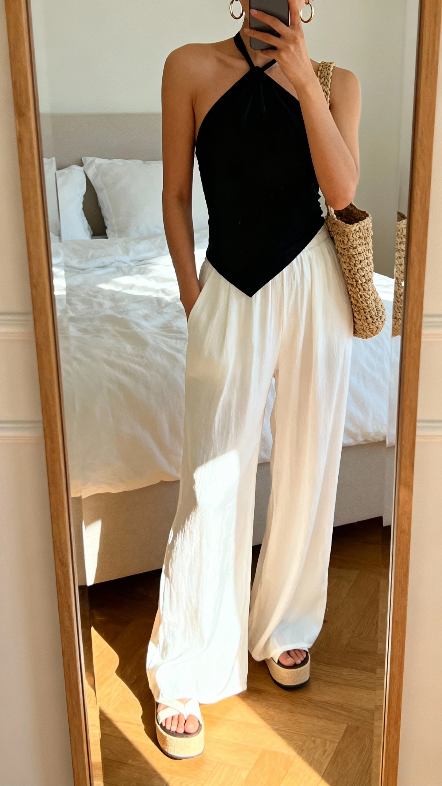 iPhone mirror selfie of a woman in flowing white palazzo pants with a chic black halter top, platform sandals, hoop earrings, and a woven shoulder bag, face not visible, cropped at neck, bedroom mirror, sunny morning light, iPhone photo quality.