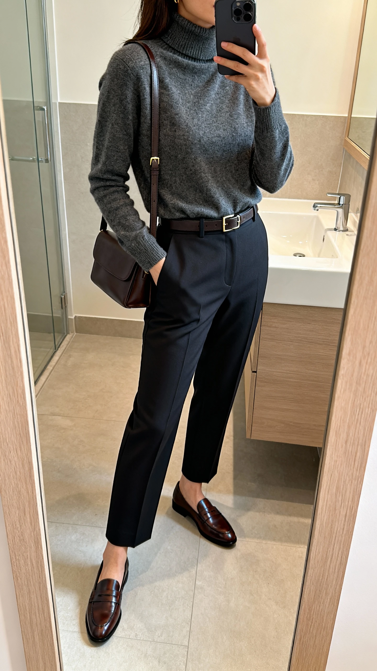 iPhone mirror selfie of a woman in pressed tailored trousers with a cashmere turtleneck, sleek leather loafers, slim belt, and a simple leather shoulder bag, face not visible, side angle in bathroom mirror, soft indoor lighting, iPhone photo quality.