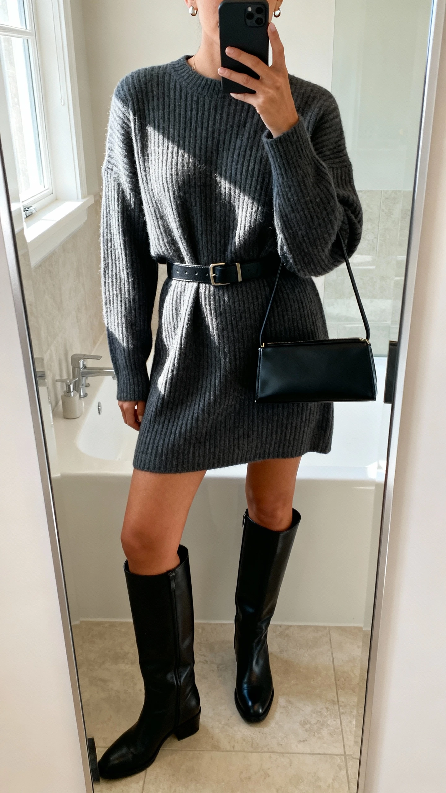 iPhone mirror selfie of a woman wearing a charcoal ribbed sweater dress with a waist belt, black knee-high leather boots, slim black top-handle bag, simple stud earrings, face not visible, cropped at shoulders, bathroom mirror, soft window light, iPhone photo quality