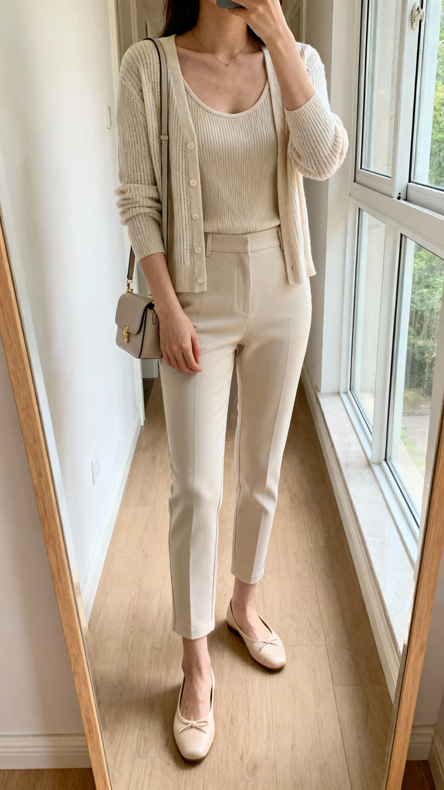 iPhone mirror selfie of a woman wearing a chic cardigan set (fine-knit cardigan and matching shell) with slim ankle pants, ballet flats, and a small shoulder bag, face not visible, cropped at shoulders, full-length hallway mirror, natural window light, iPhone photo quality