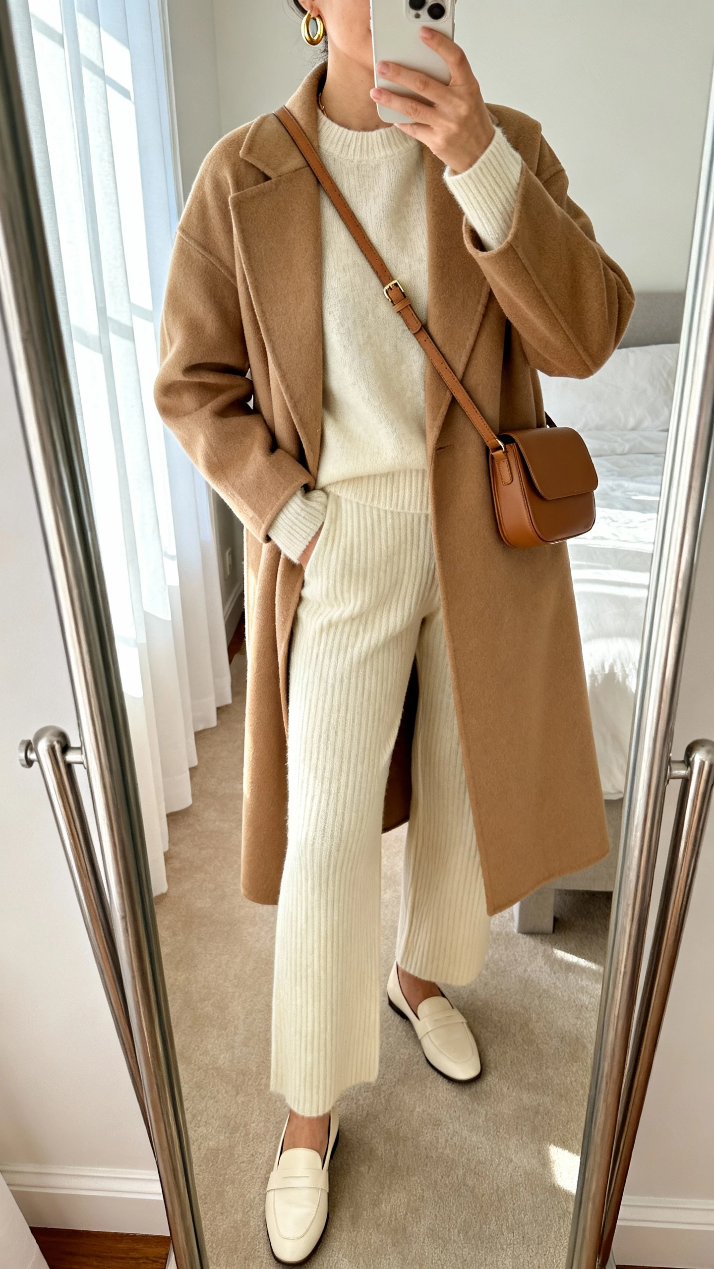 iPhone mirror selfie of a woman wearing a creamy cashmere matching set (crewneck sweater and knit trousers) layered with a tailored camel coat, delicate gold hoop earrings, minimalist leather crossbody, and cream loafers, face not visible, cropped at neck, bedroom mirror, soft morning window light, iPhone photo quality.