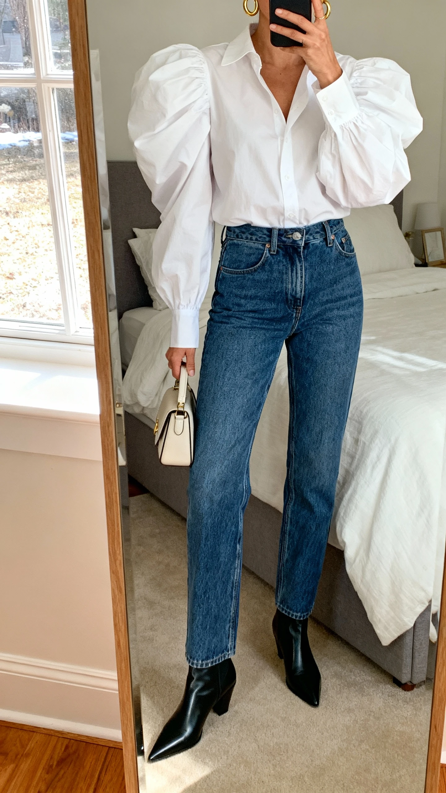 iPhone mirror selfie of a woman wearing a dramatic puff-sleeve blouse in crisp white paired with high-waisted straight jeans and pointed ankle boots, simple gold hoops and a small structured handbag, face not visible, cropped at neck, bedroom mirror, natural window light, iPhone photo quality.
