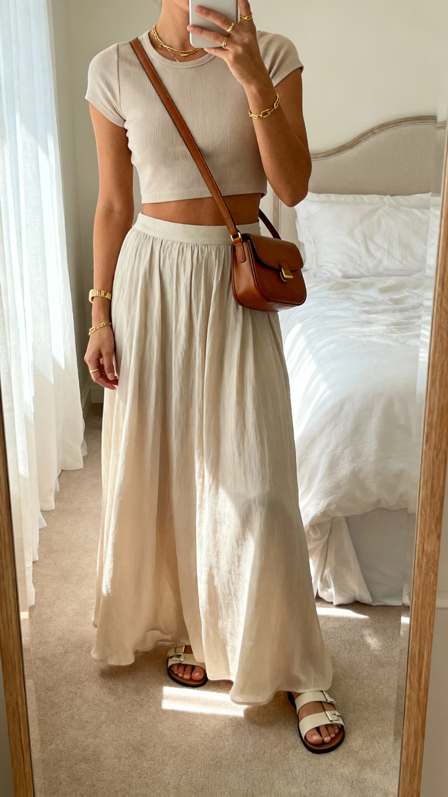 iPhone mirror selfie of a woman wearing a flowy maxi skirt with a fitted crop top, sleek sandals, delicate gold jewelry, and a small leather shoulder bag for quiet-luxury vibes, face not visible, bedroom mirror, soft morning light, iPhone photo quality.
