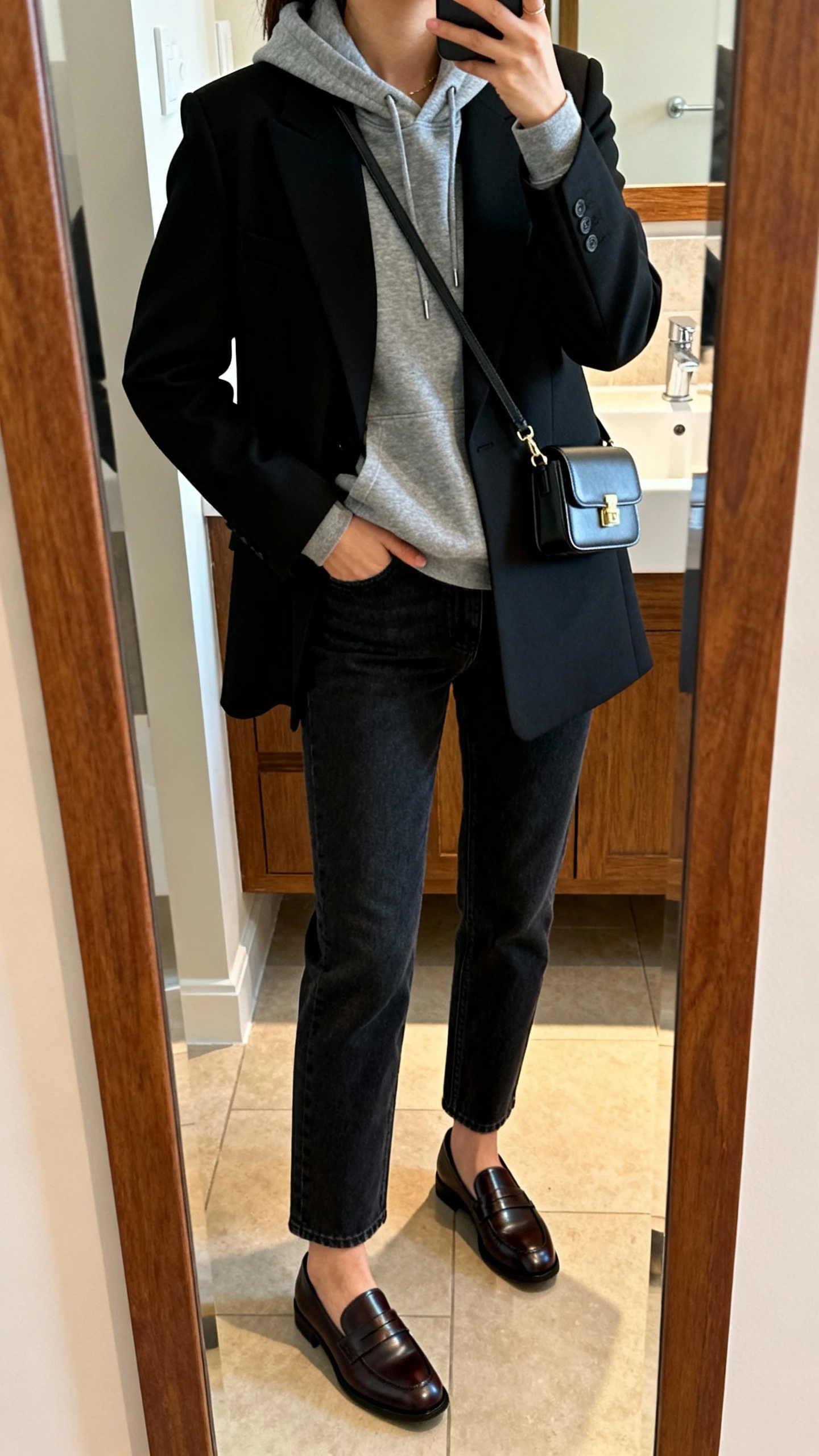 iPhone mirror selfie of a woman wearing a gray hoodie layered under a tailored black blazer, dark straight-leg jeans, sleek leather loafers, mini crossbody bag, face not visible, cropped at neck, bathroom mirror, indoor warm lighting, iPhone photo quality