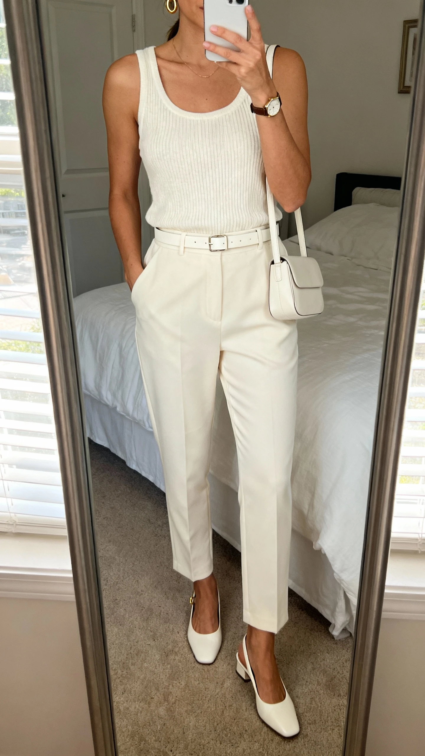 iPhone mirror selfie of a woman wearing a monochrome set: cream knit tank, matching cream tailored trousers, cream minimalist belt, cream slingback flats, small cream shoulder bag, delicate gold hoops and watch, face not visible, cropped at neck, bedroom mirror with natural morning window light, iPhone photo quality.