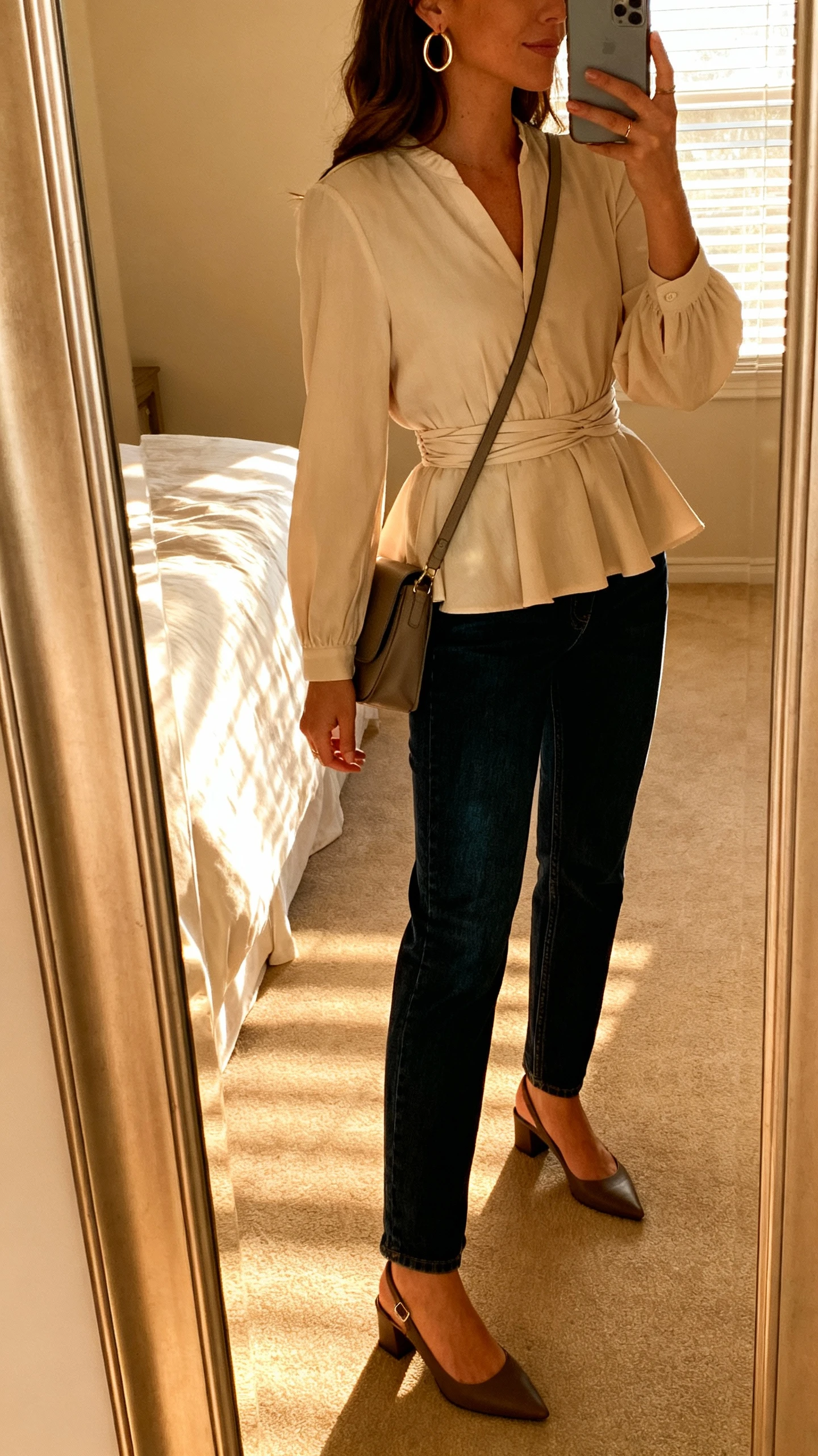 iPhone mirror selfie of a woman wearing a peplum blouse that cinches at the waist with straight-leg dark denim and slingback heels, hoop earrings and a crossbody bag, face not visible, side angle, bedroom mirror, afternoon window light, iPhone photo quality.