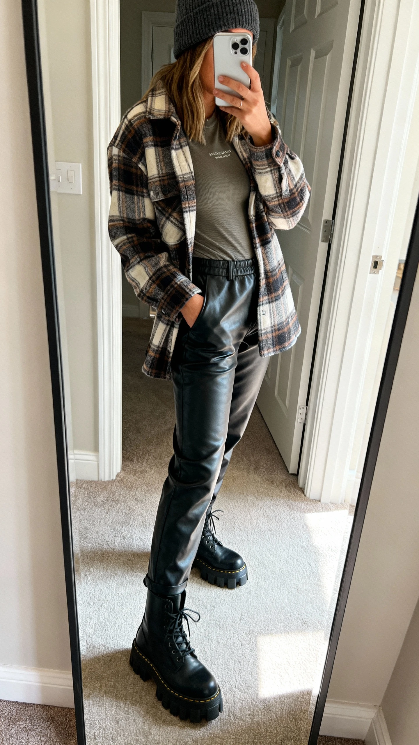 iPhone mirror selfie of a woman wearing a plaid shacket over a fitted tee, faux leather pants, lug-sole boots, and a beanie, face not visible, side angle, entryway mirror, natural daylight, iPhone photo quality.