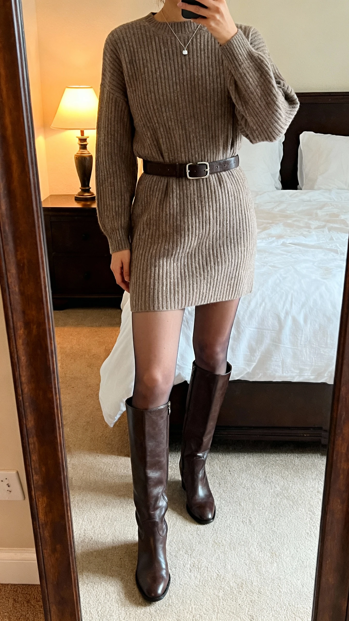 iPhone mirror selfie of a woman wearing a ribbed sweater dress with tall knee-high leather boots, belt at the waist, sheer tights, and a minimal pendant necklace; face not visible, cropped at shoulders, bedroom mirror, warm indoor lamp light, iPhone photo quality.