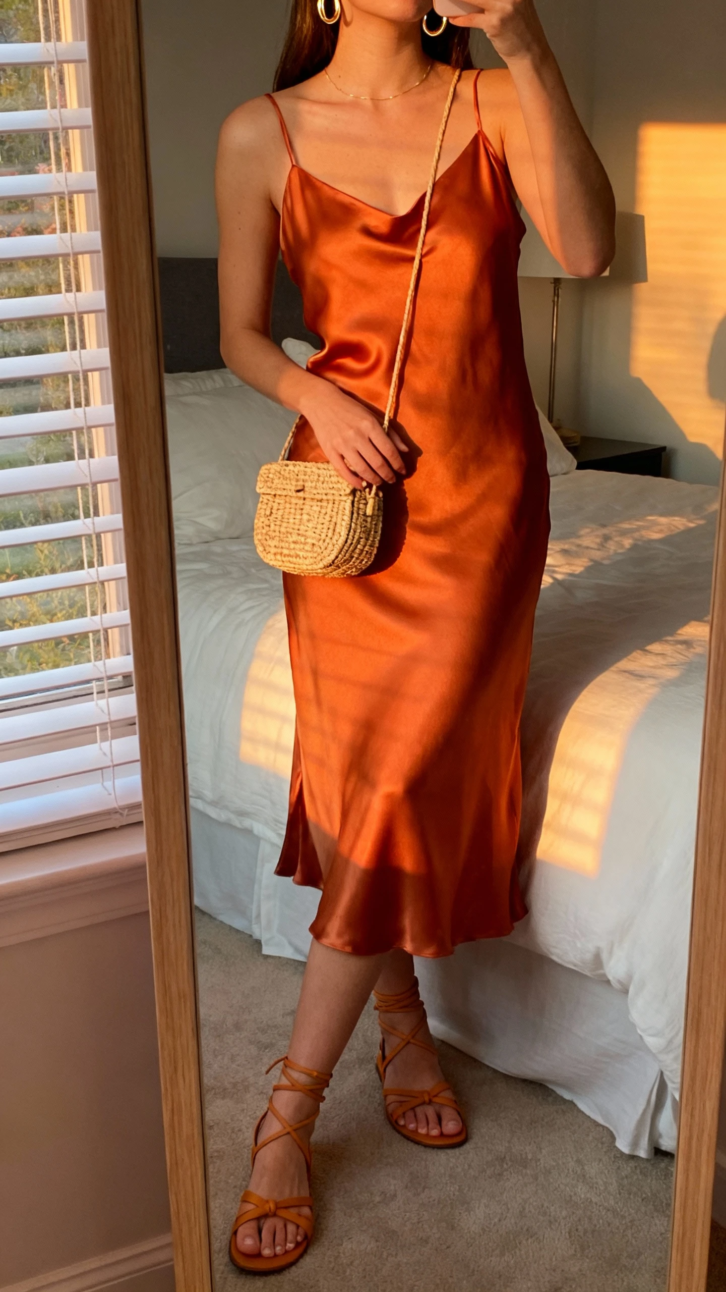 iPhone mirror selfie of a woman wearing a satin sunset-orange slip dress styled with delicate gold hoops, strappy tan sandals, and a raffia mini bag, face not visible, cropped at neck, bedroom mirror, warm evening window light, iPhone photo quality.