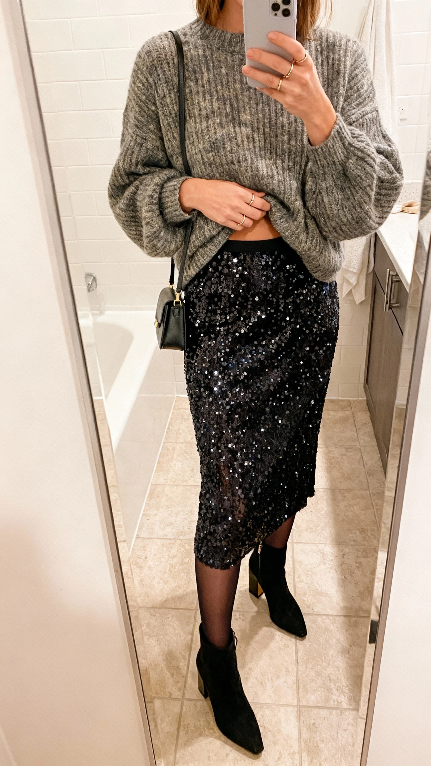 iPhone mirror selfie of a woman wearing a sequin midi skirt with a chunky knit sweater half-tucked, sheer black tights, block-heel ankle boots, small shoulder bag, and delicate rings; face not visible, cropped at neck, bathroom mirror, warm indoor lighting, iPhone photo quality.