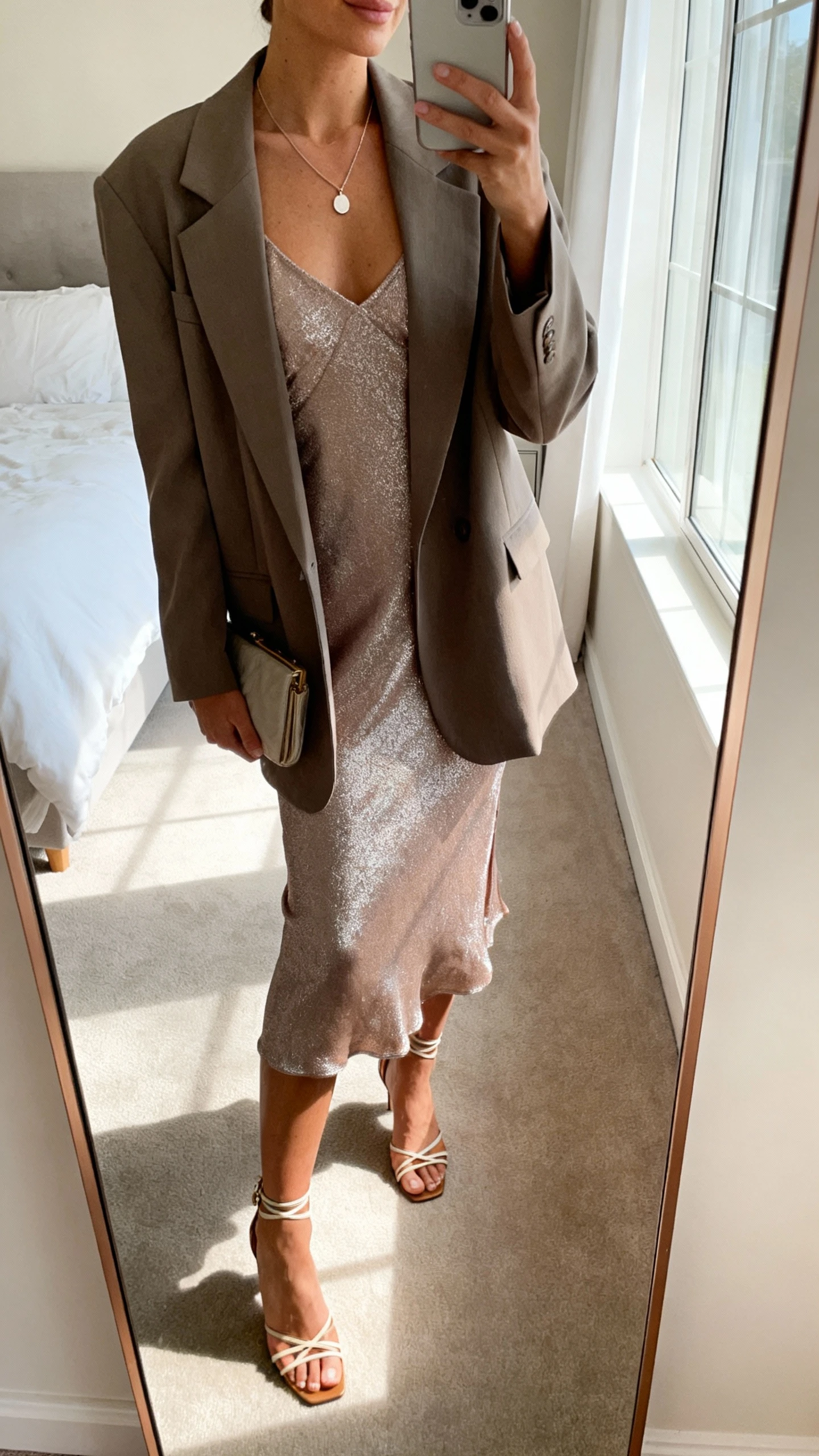 iPhone mirror selfie of a woman wearing a shimmer slip dress with an oversized blazer, strappy heels, delicate pendant necklace, and a small clutch, face not visible, cropped at neck, bedroom mirror, soft morning window light, iPhone photo quality.