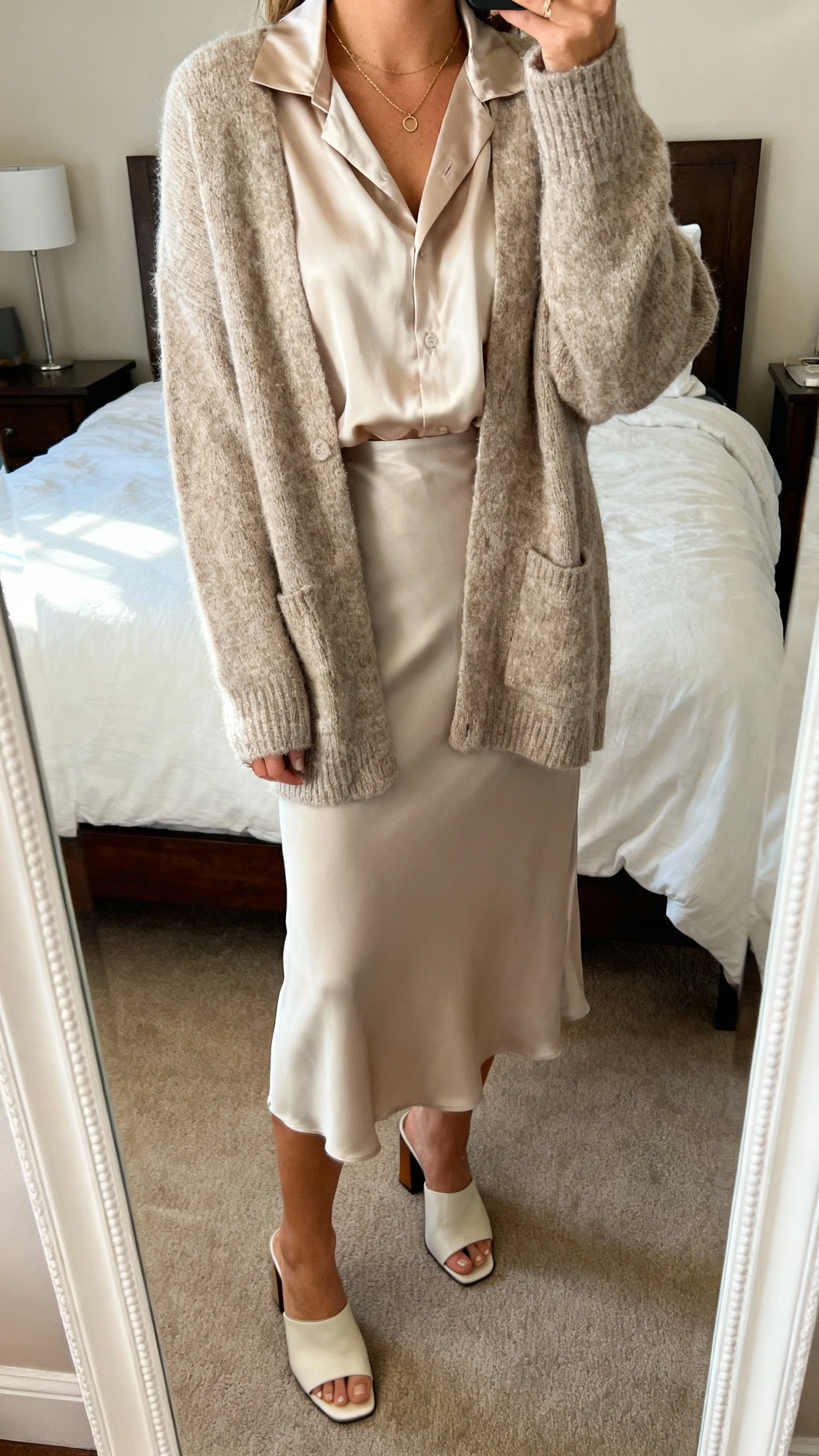 iPhone mirror selfie of a woman wearing a silk blouse under a cozy cardigan with a midi slip skirt, delicate necklace, and heeled mules, face not visible, cropped at shoulders, bedroom mirror, soft natural light, iPhone photo quality.