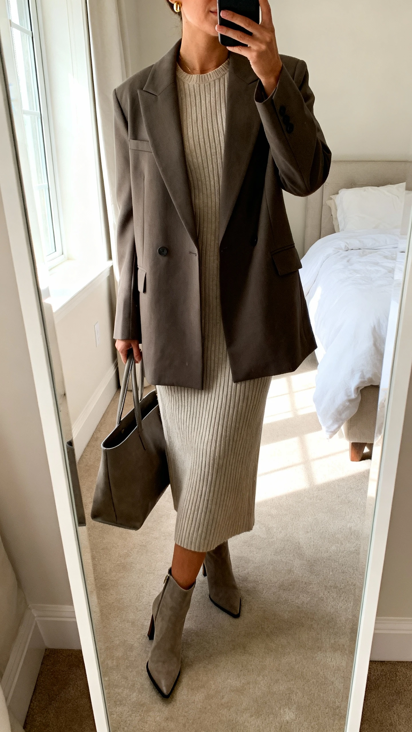 iPhone mirror selfie of a woman wearing a tailored power coat layered over a ribbed knit midi dress with heeled ankle boots, structured tote, and minimal gold studs, face not visible, cropped at neck, bedroom mirror, morning window light, iPhone photo quality.