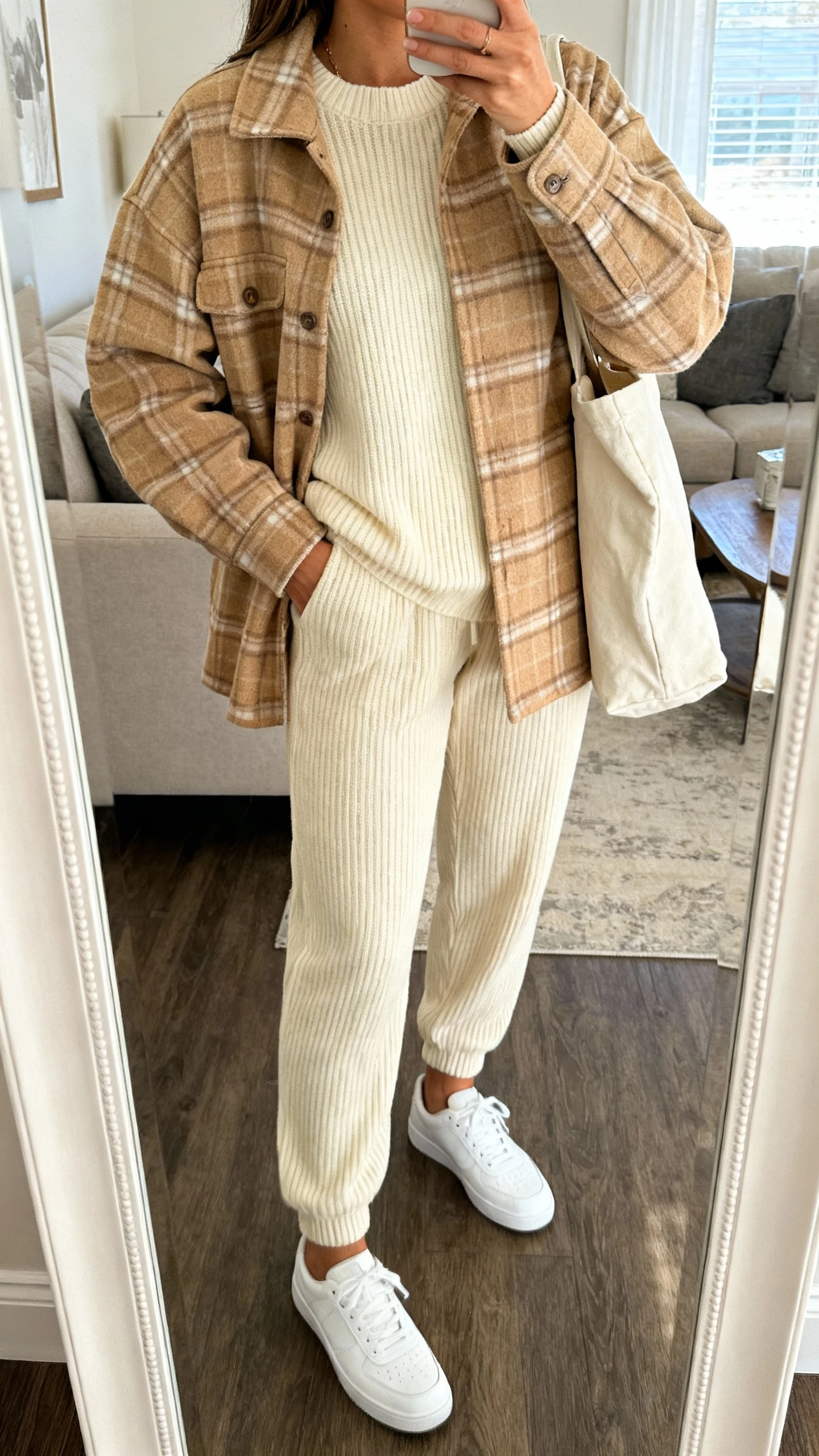 iPhone mirror selfie of a woman wearing a tan plaid shacket over a matching cream knit set (ribbed sweater and knit joggers), white low-top sneakers, soft tote bag, face not visible, cropped at shoulders, living-room mirror, natural daylight, iPhone photo quality