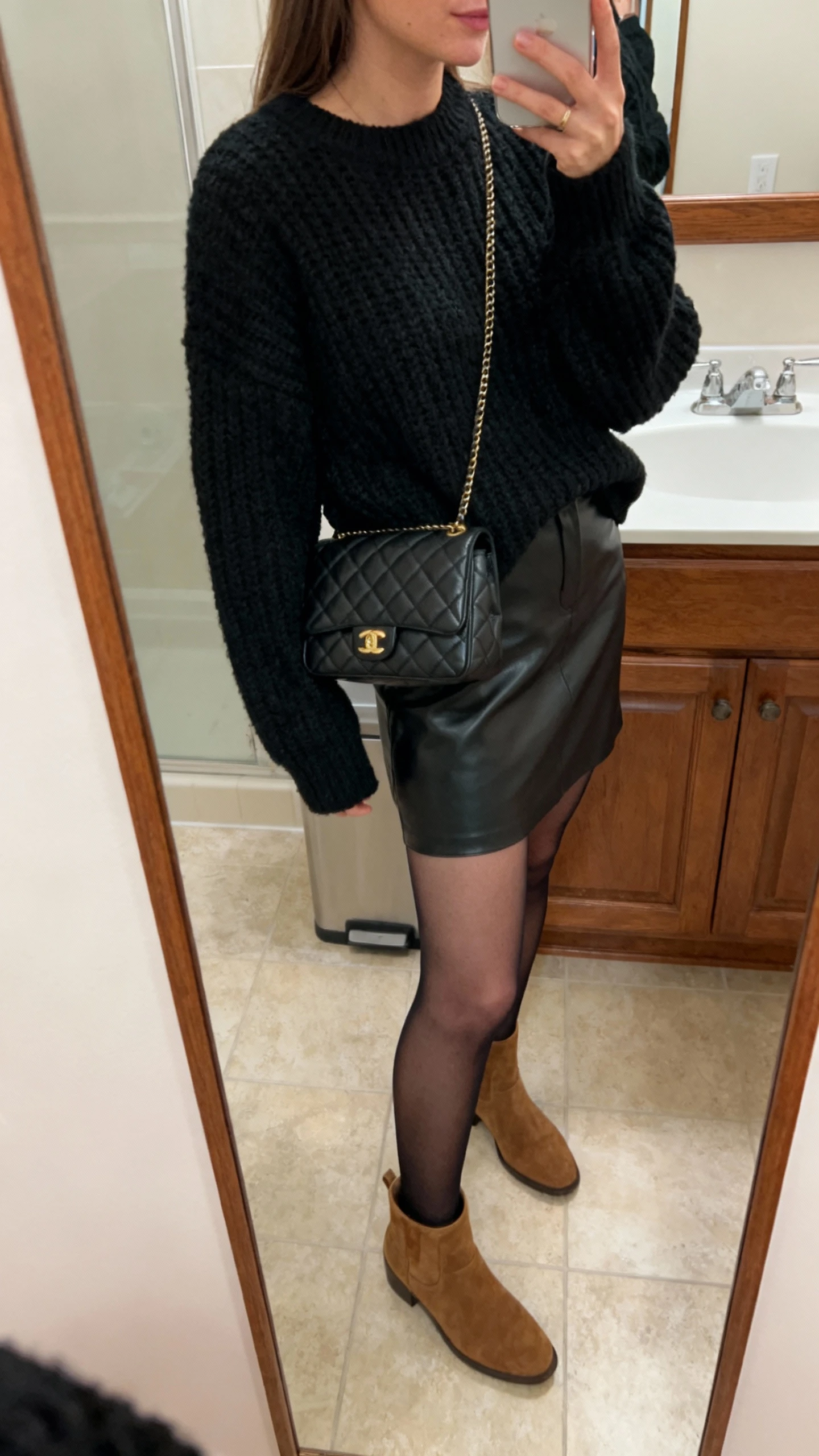 iPhone mirror selfie of a woman wearing an all-black monochrome look: textured knit sweater, faux leather skirt, sheer tights, suede ankle boots, and a quilted bag, face not visible, bathroom mirror, side angle, indoor lighting, iPhone photo quality.
