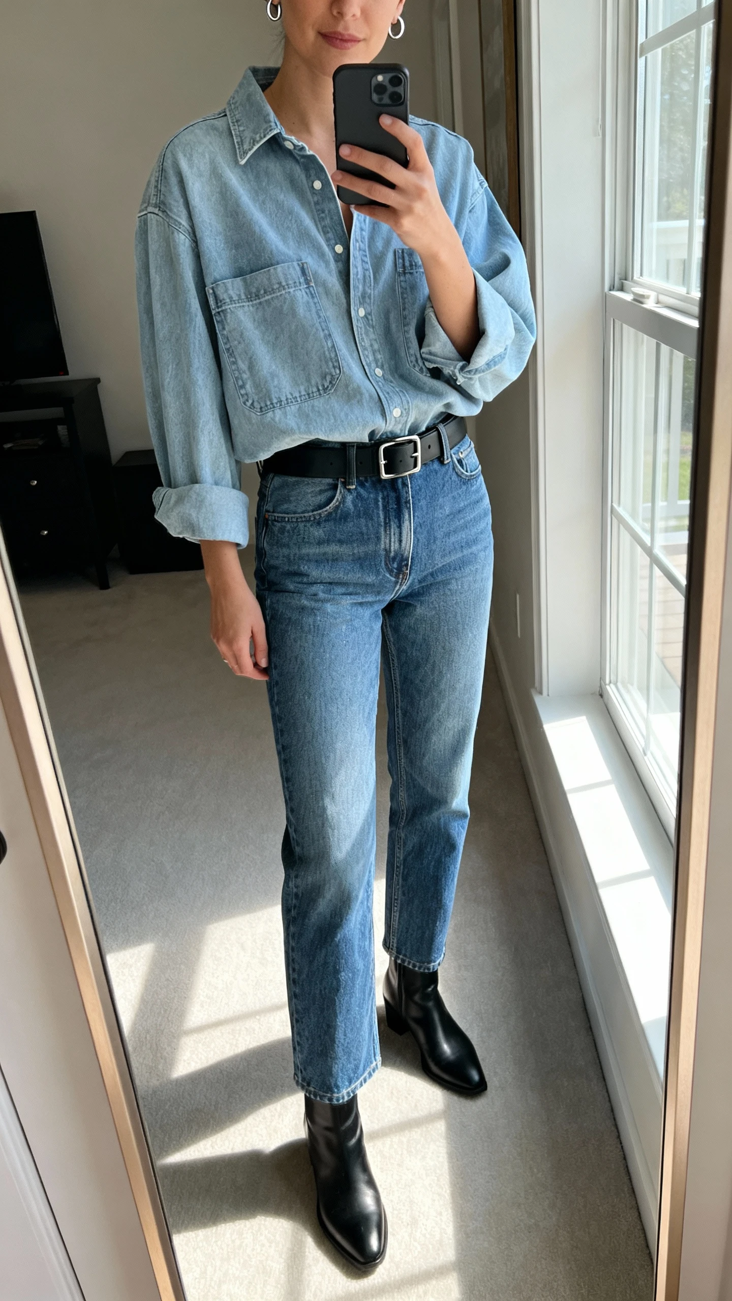 iPhone mirror selfie of a woman wearing minimal denim-on-denim: light-wash oversized denim shirt tucked into mid-wash straight jeans, black leather belt, black ankle boots, tiny silver hoops, face not visible, cropped at shoulders, studio-apartment mirror with natural window light, iPhone photo quality.
