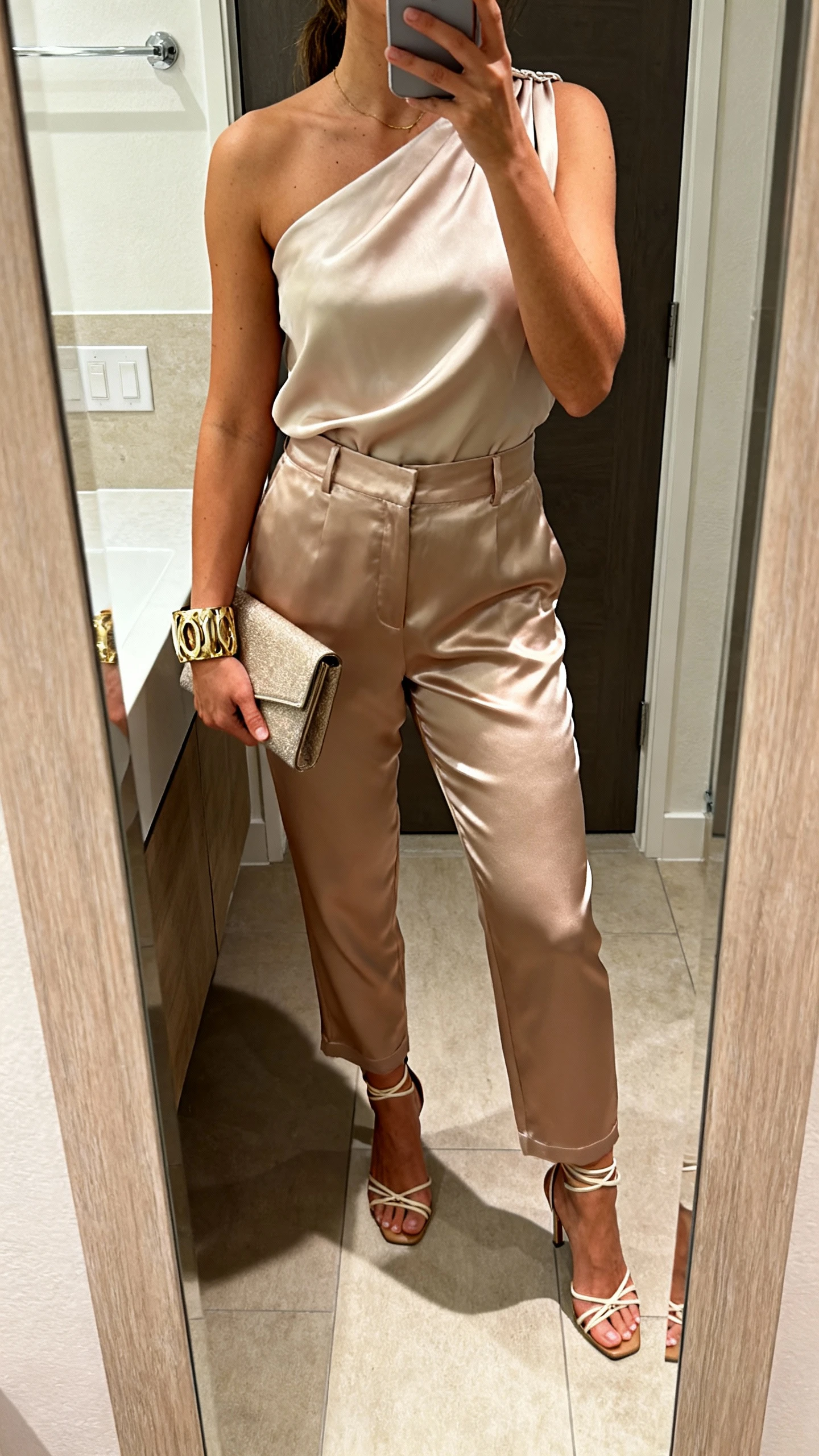 iPhone mirror selfie of a woman wearing satin pants with a one-shoulder top, strappy heels, statement cuff bracelet, and a clutch, face not visible, cropped at neck, bathroom mirror, warm indoor lighting, iPhone photo quality.
