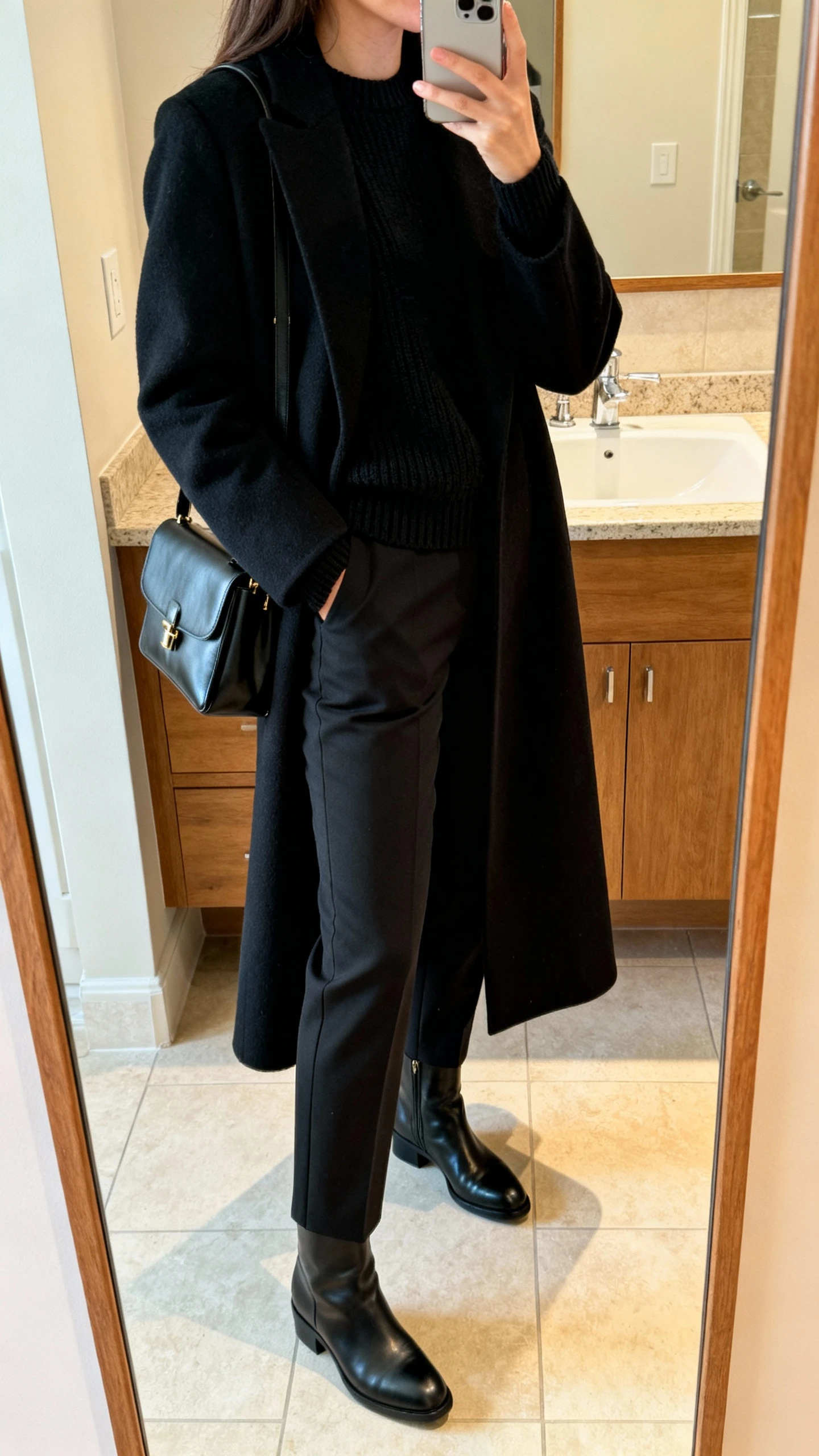 iPhone side-angle mirror selfie of a woman in all-black tonal layers: long coat, crewneck knit, tailored trousers, leather boots, and a structured bag, face not visible, bathroom mirror, warm indoor lighting, iPhone photo quality.