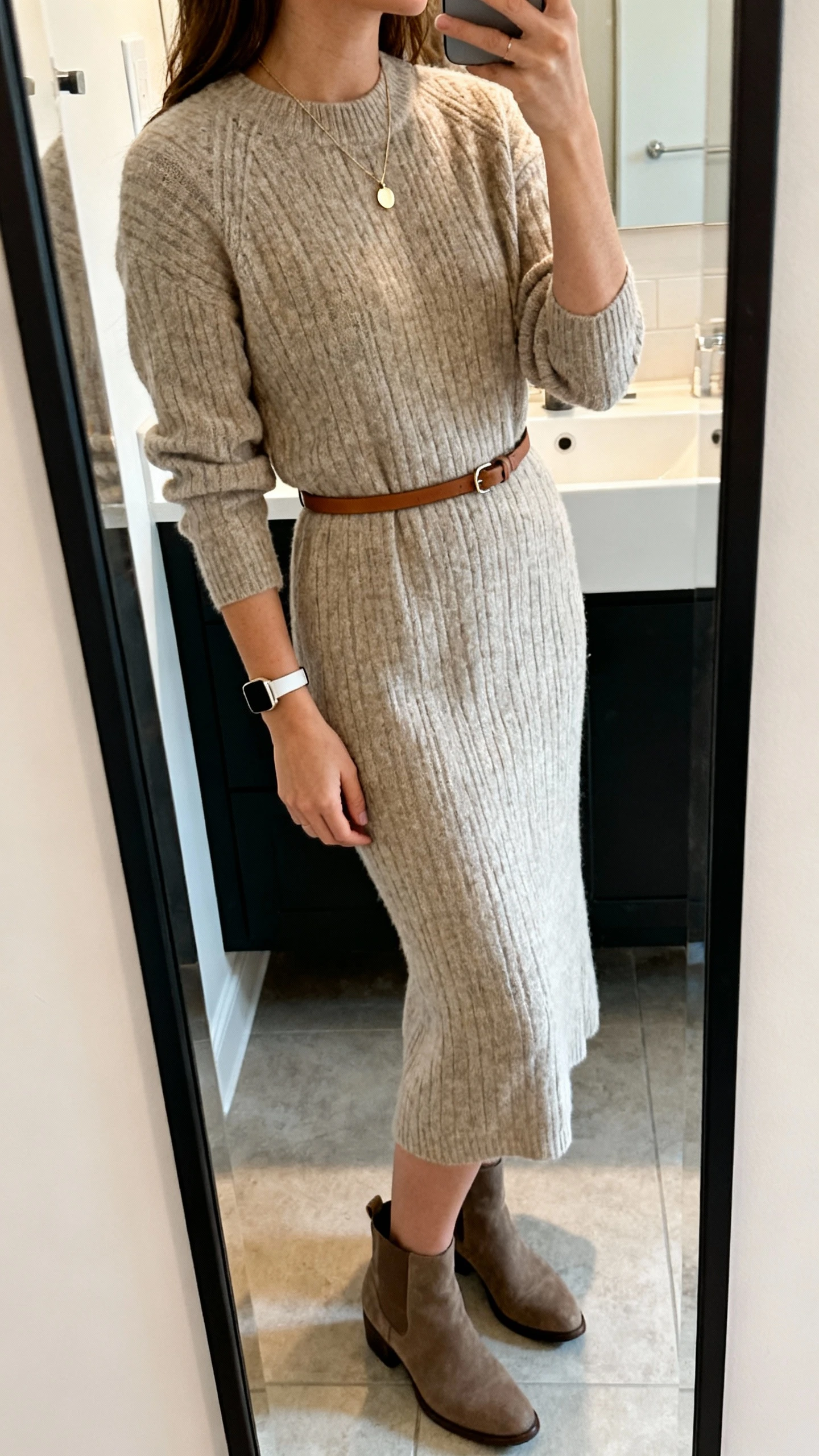 iPhone side-angle mirror selfie of a woman in an elevated knit midi dress with a thin leather belt, ankle boots, delicate pendant necklace, and a slim watch, face not visible, bathroom mirror, soft indoor lighting, iPhone photo quality