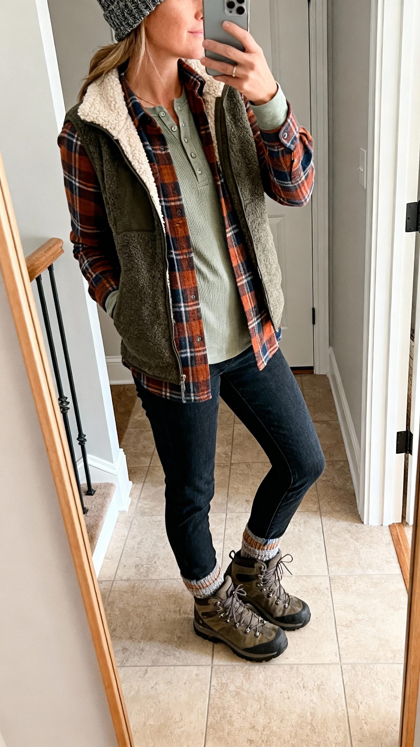 iPhone side-angle mirror selfie of a woman in cabin-cool flannel layered over a thermal henley, fleece-lined vest, straight-leg dark jeans, hiking-inspired boots, wool socks peeking out, and a knit beanie; face not visible, entryway mirror, afternoon natural light, iPhone photo quality.