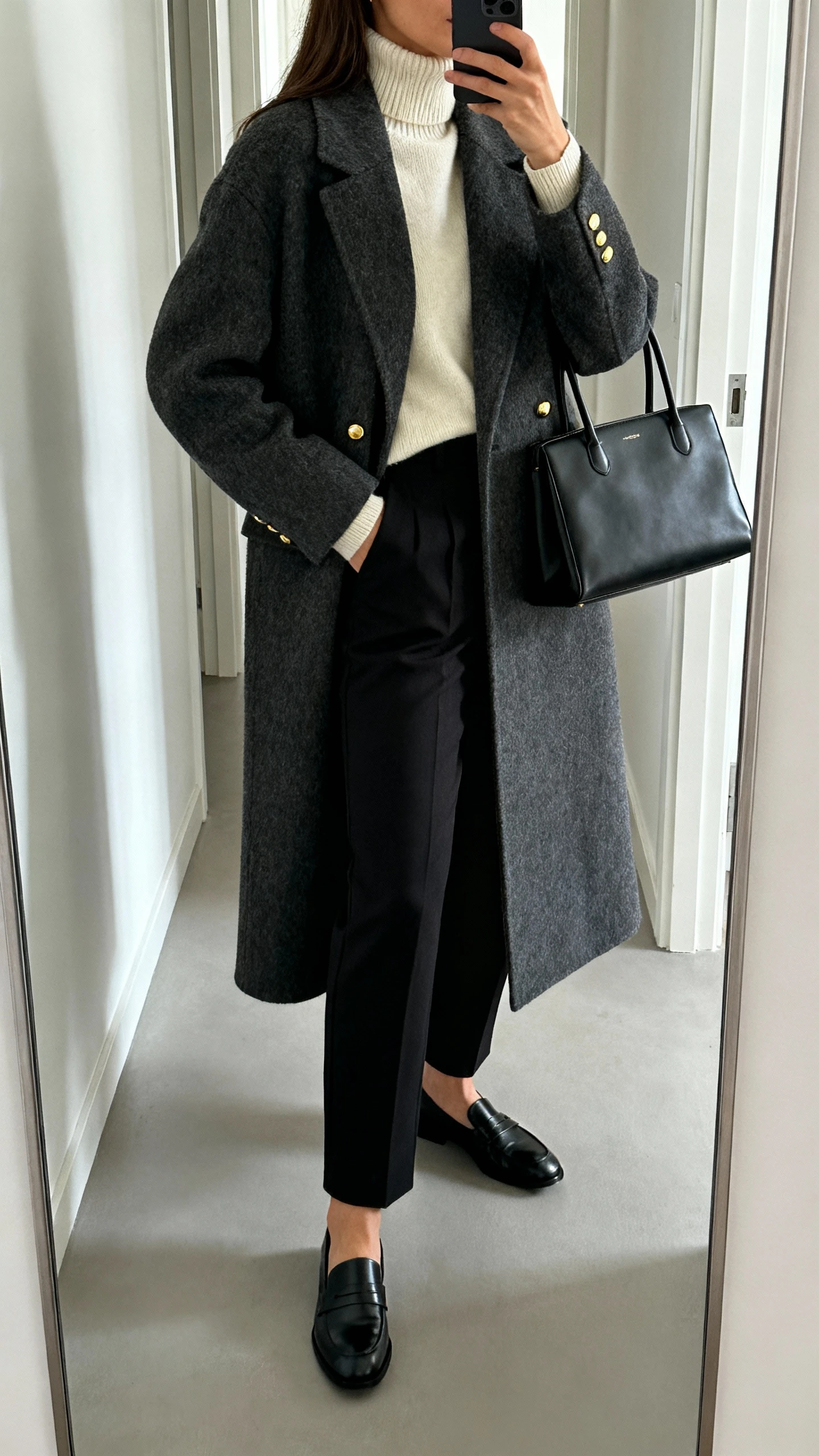 iPhone side-angle mirror selfie of a woman in Nordic minimalist layers: charcoal wool coat, cream merino turtleneck, tailored black trousers, black leather loafers, structured black tote, and simple gold studs; face not visible, hallway mirror, cool natural light, iPhone photo quality.