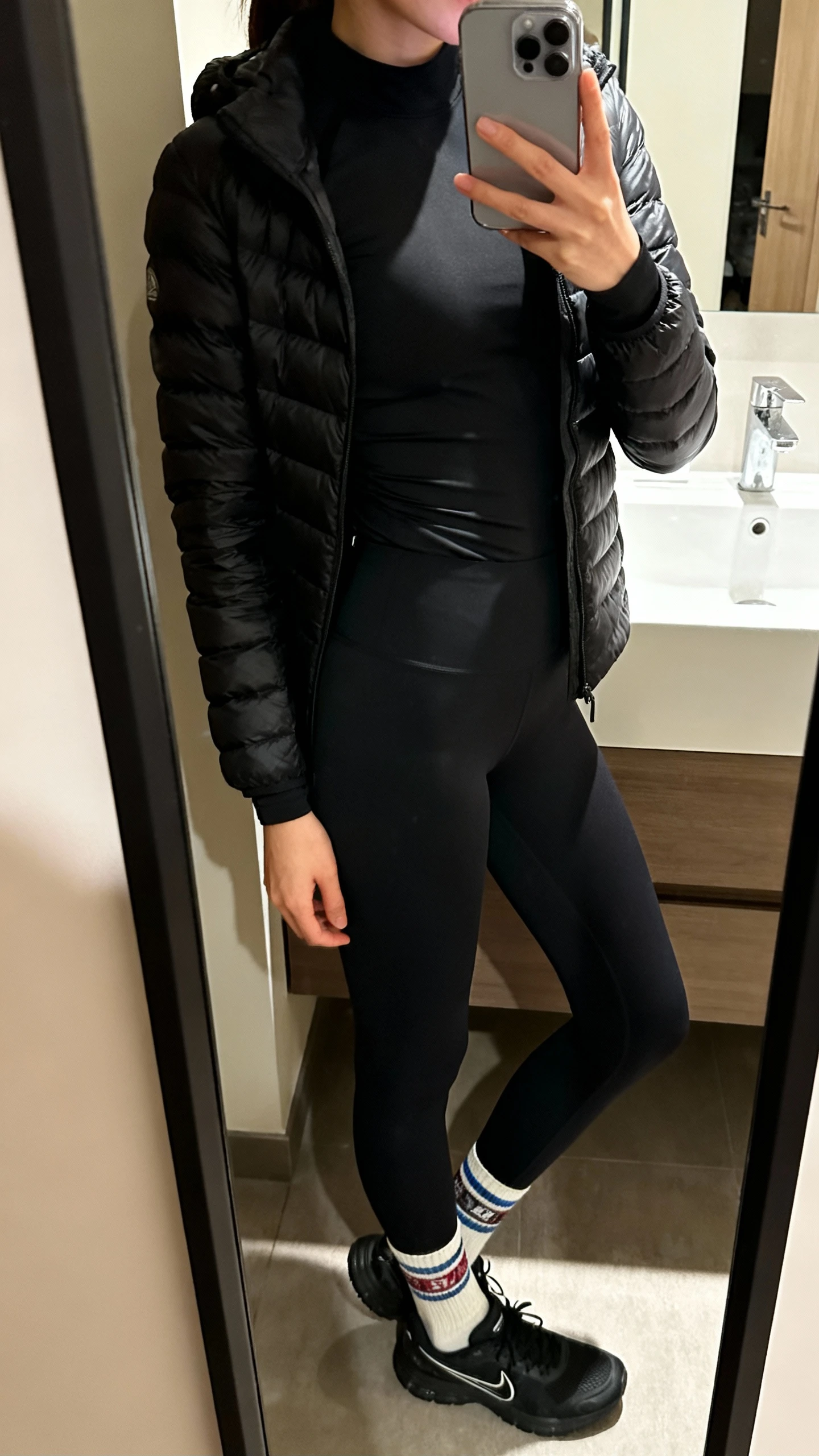 iPhone side-angle mirror selfie of a woman wearing black leggings, a sleek black puffer jacket, a fitted long-sleeve base layer, sporty crew socks, and black running sneakers, face not visible, bathroom mirror with indoor lighting, iPhone photo quality.