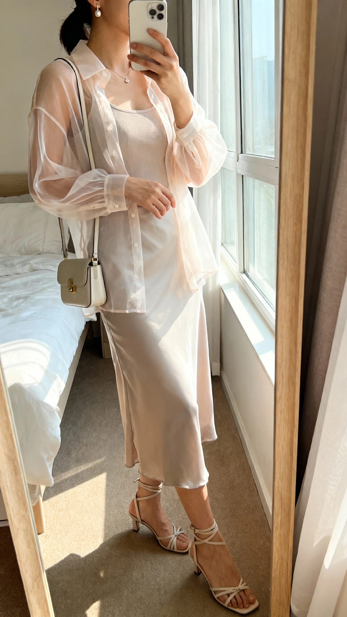 iPhone side-angle mirror selfie of a woman wearing a sheer organza blouse over a fitted camisole, styled with a slip midi skirt and strappy heels, petite shoulder bag and dainty earrings, face not visible, cropped at shoulders, bedroom mirror, morning window light, iPhone photo quality.