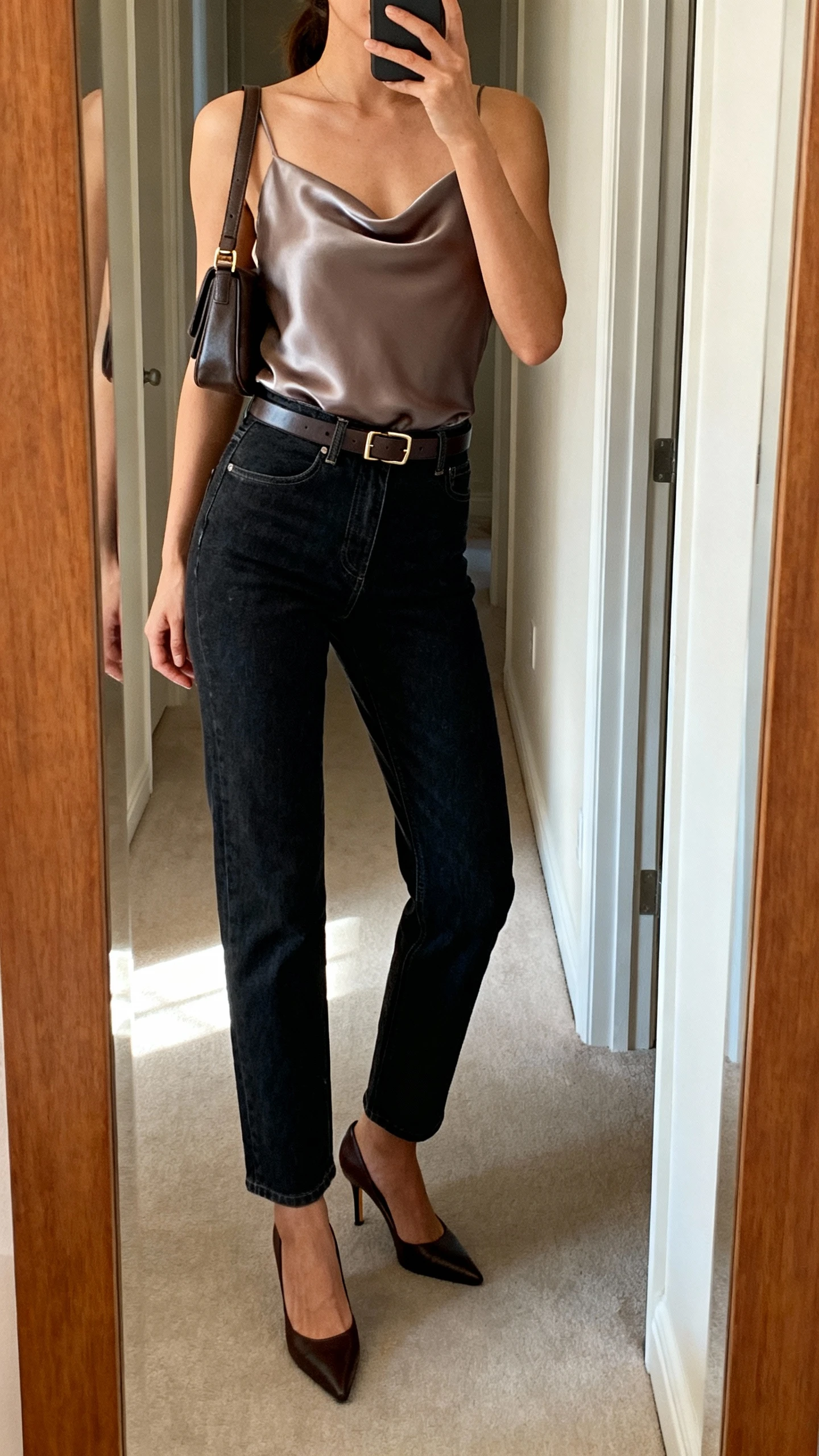 iPhone side-angle mirror selfie of a woman wearing straight-leg dark-wash jeans, a silky camisole, classic heels, a slim belt, and a structured shoulder bag, face not visible, cropped at shoulders, full-length mirror in hallway, natural afternoon light, iPhone photo quality.