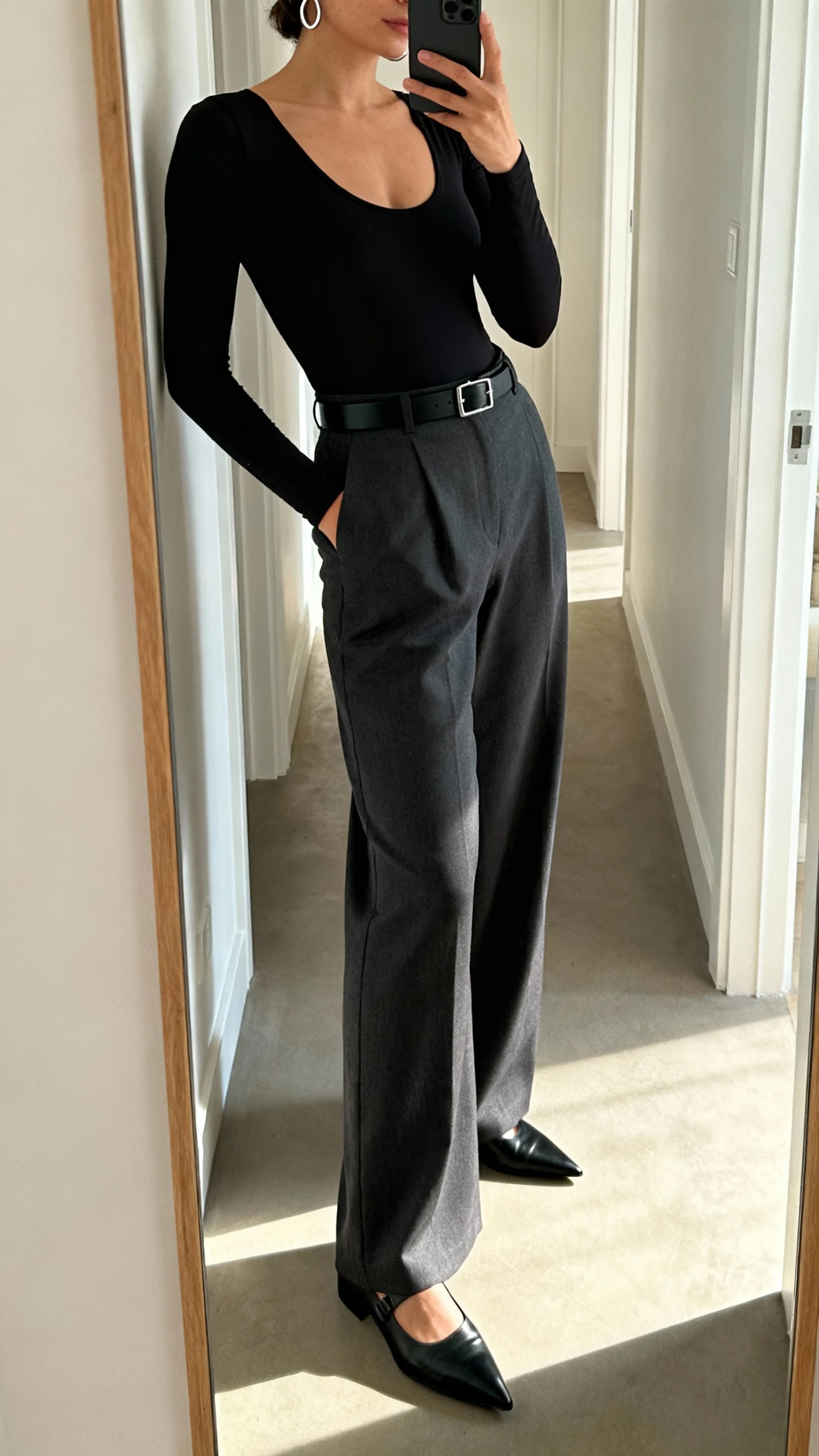 iPhone side-angle mirror selfie of a woman wearing a sleek black bodysuit with high-waisted charcoal wide-leg trousers, pointed black loafers, slim black belt, simple silver hoops, face not visible, cropped at shoulders, full-length hallway mirror, natural afternoon light, iPhone photo quality.