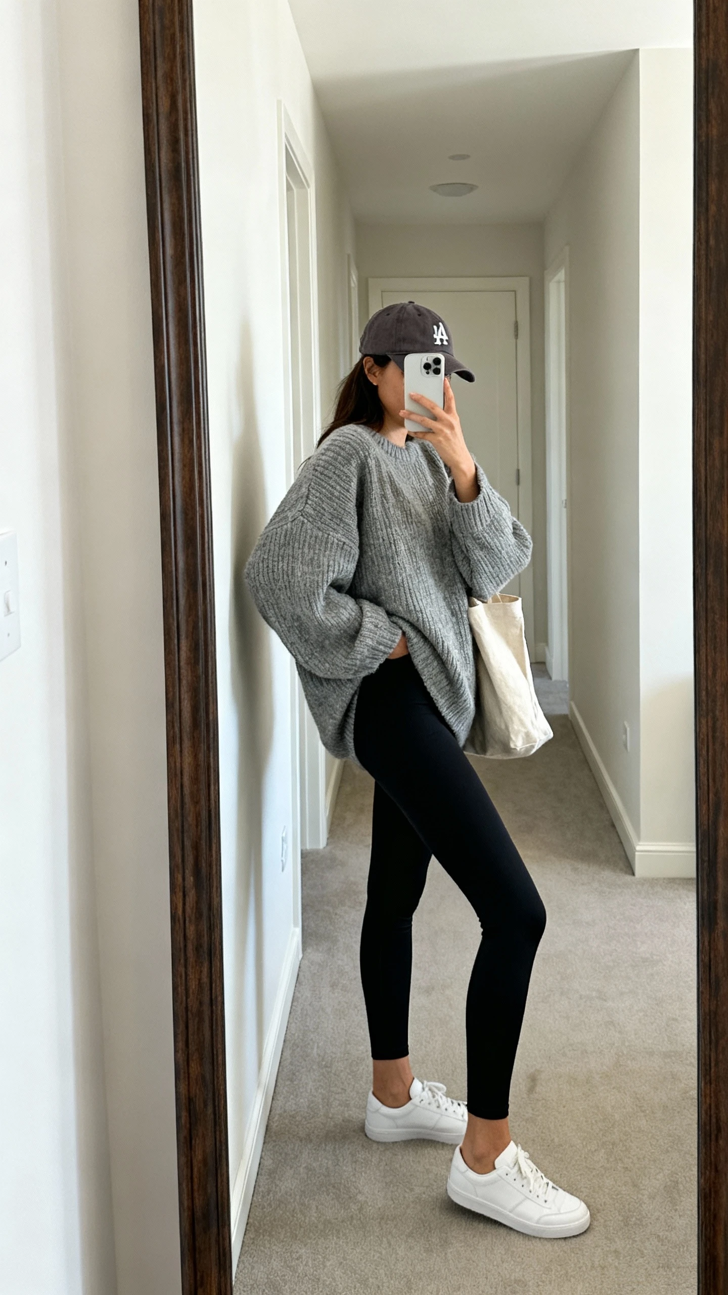 iPhone side-angle mirror selfie of a woman wearing an oversized heather gray knit sweater, black high-waist leggings, white low-top sneakers, a baseball cap, and a canvas tote, face not visible, hallway full-length mirror, natural daylight, iPhone photo quality.