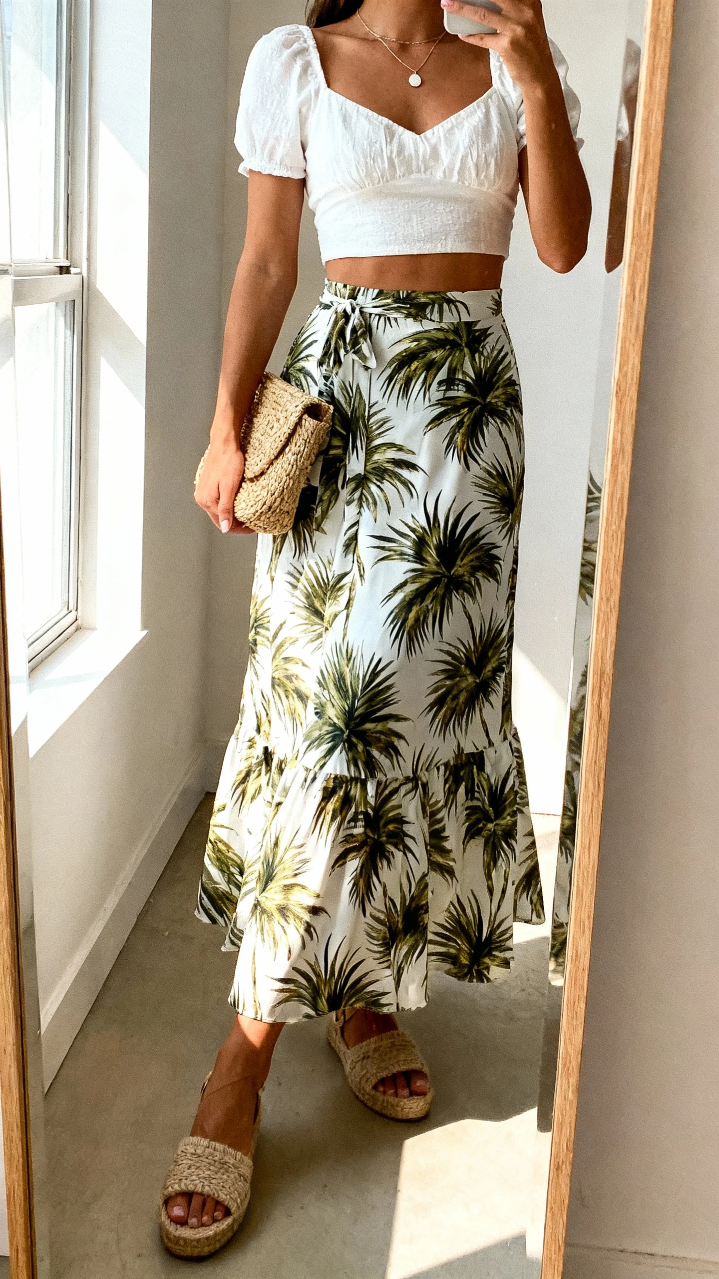 iPhone side-angle mirror selfie of a woman wearing a palm-print flowy maxi skirt with a flirty white cropped top, espadrille sandals, dainty pendant necklace, and a straw clutch, face not visible, full-length mirror, bright window light, iPhone photo quality.