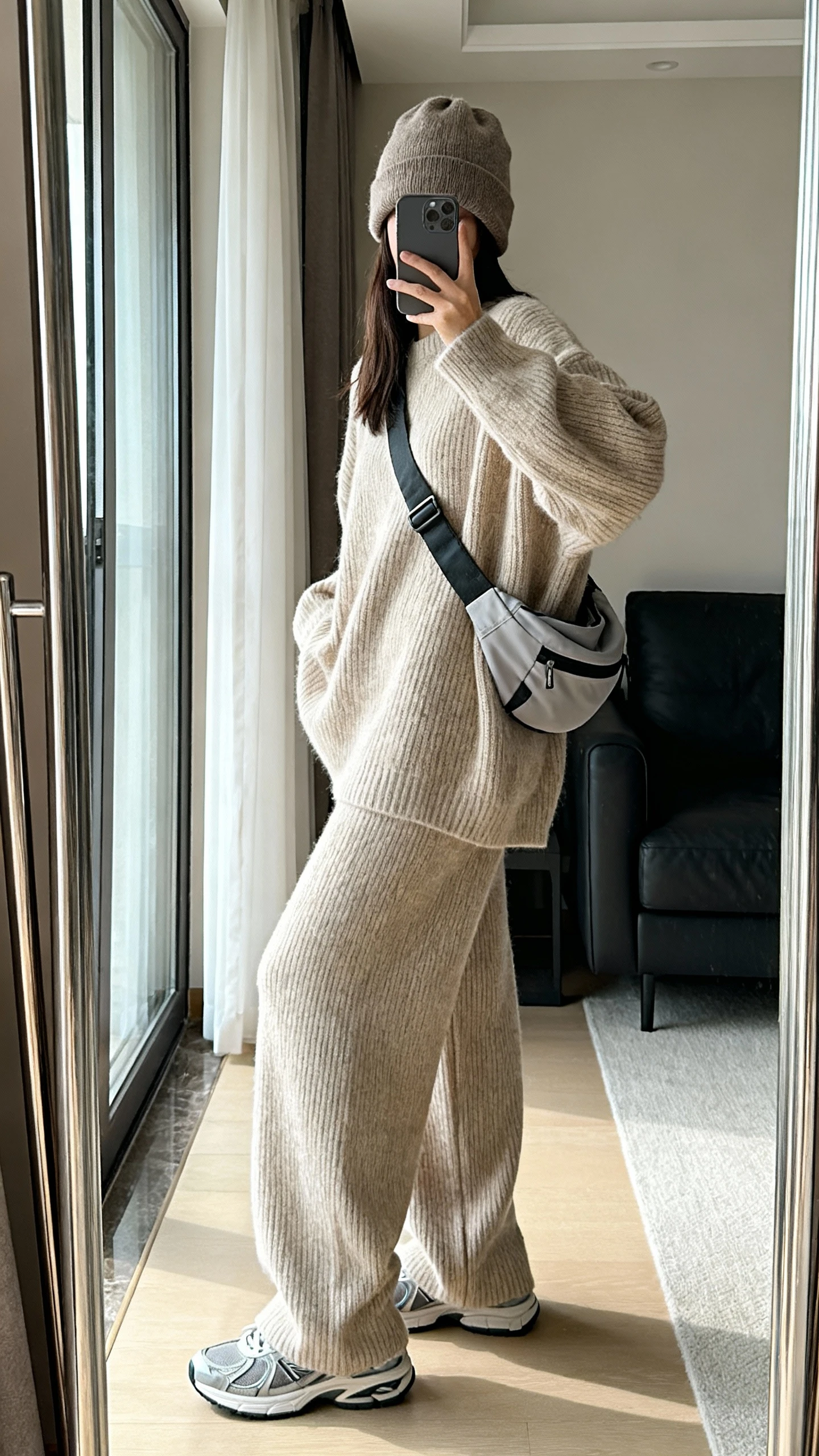 iPhone side-angle mirror selfie of a woman wearing an oversized oatmeal knit sweater with matching wide-leg knit pants, sleek trainers, a slouchy beanie, and a sporty crossbody, face not visible, living room mirror, soft natural window light, iPhone photo quality.