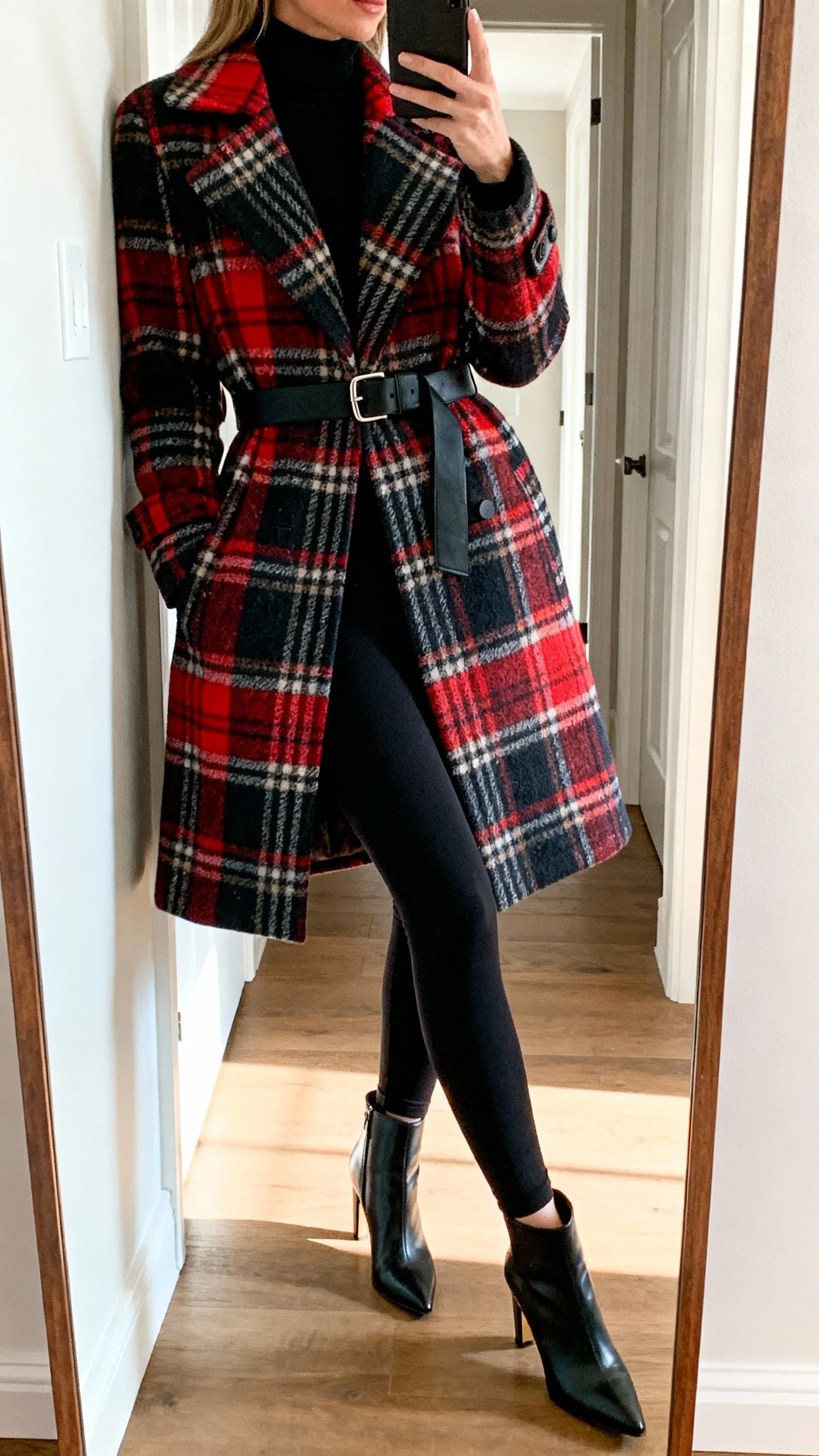 iPhone side-angle mirror selfie of a woman wearing black leggings, a simple black turtleneck, and a bold statement coat in plaid with belt, paired with sleek heeled ankle boots, face not visible, entryway full-length mirror with natural daylight, iPhone photo quality.