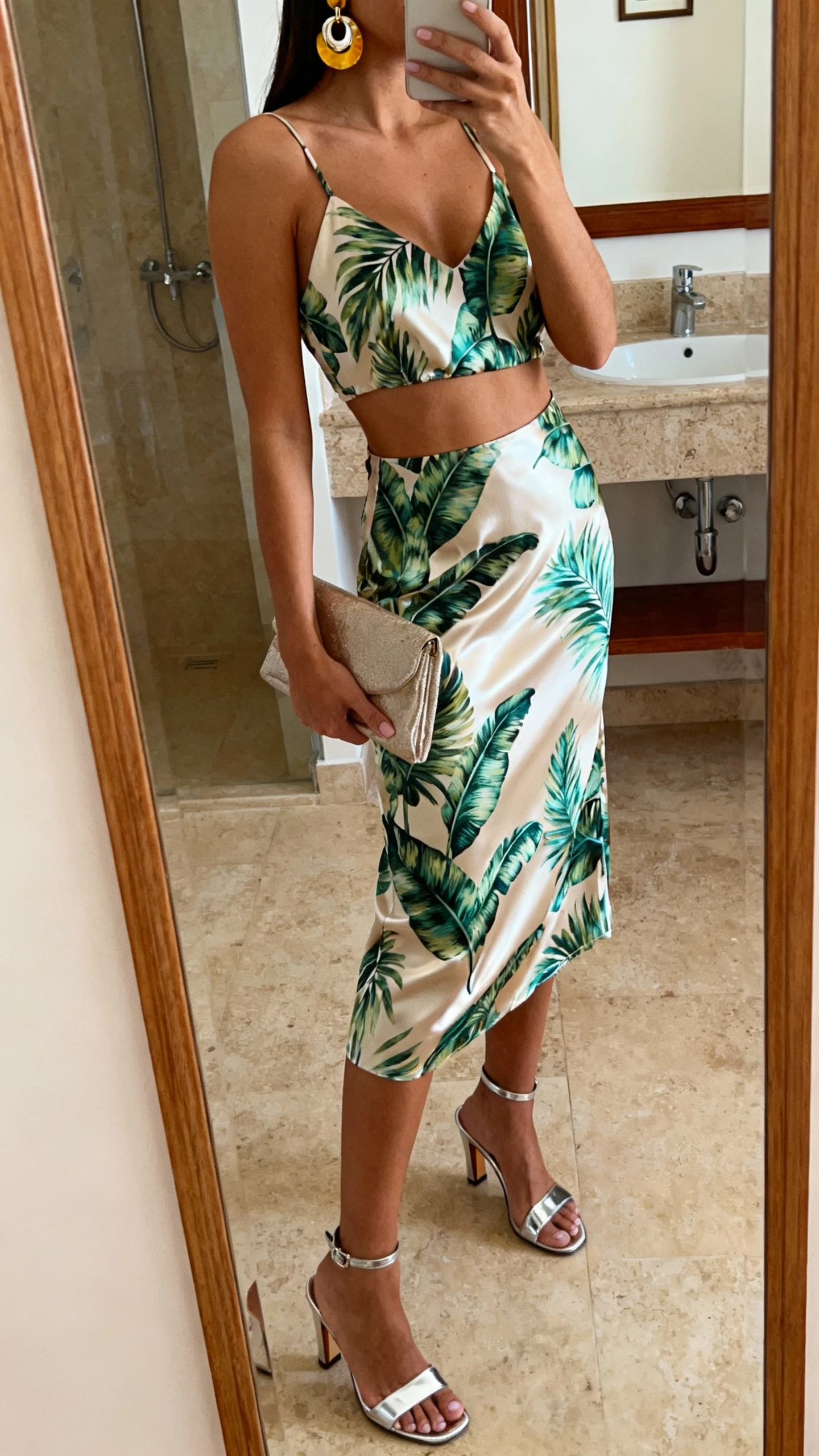 iPhone side-angle mirror selfie of a woman wearing a satin co-ord set (tropical-print camisole and matching midi skirt) with metallic heeled sandals, bold earrings, and a clutch, face not visible, bathroom mirror, warm indoor lighting, iPhone photo quality.