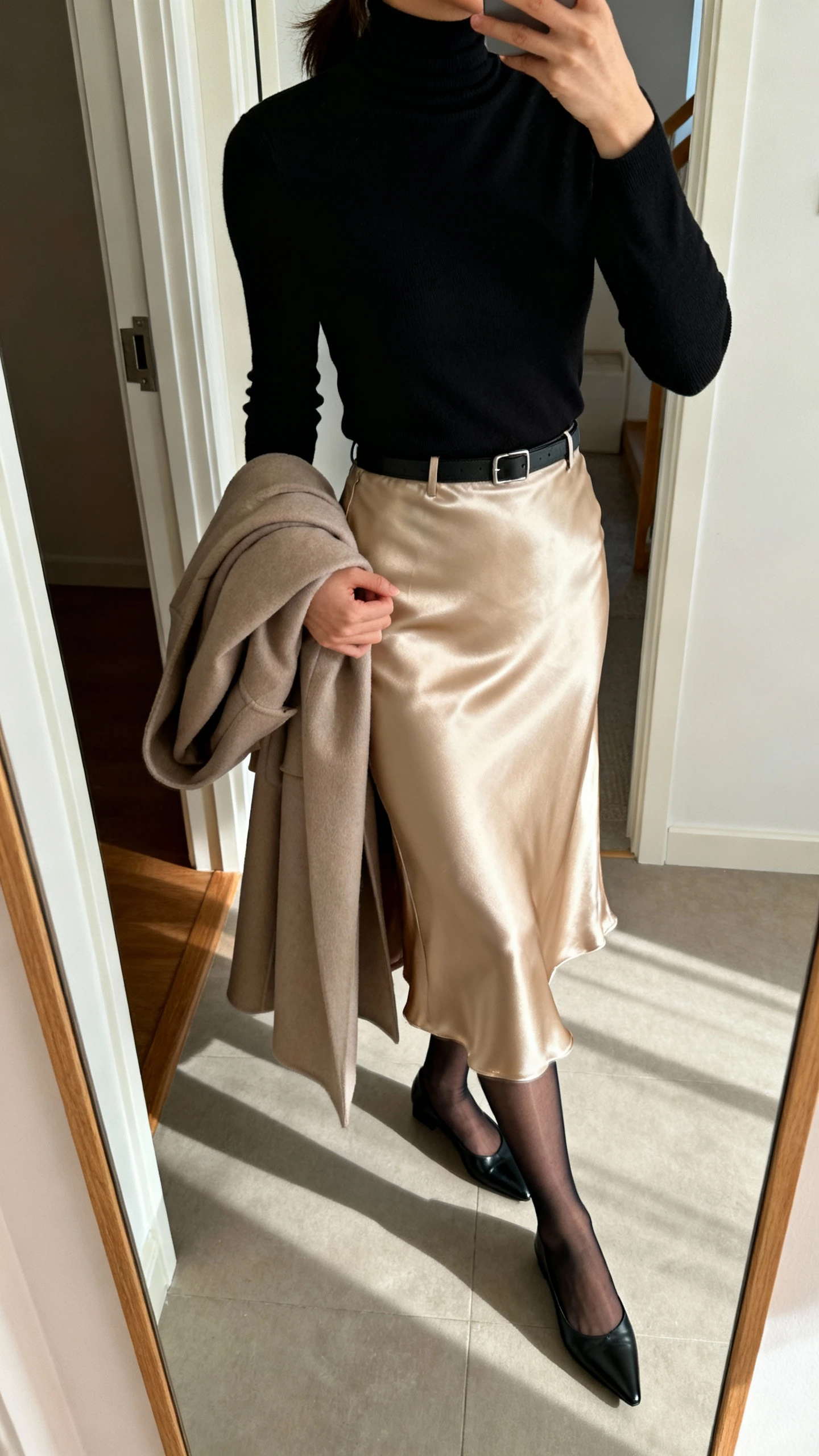 iPhone side view of a woman wearing a fitted black turtleneck tucked into a champagne satin slip skirt, sheer black tights, pointed black flats, lightweight longline coat draped over arm, face not visible, entryway mirror, morning light, iPhone photo quality