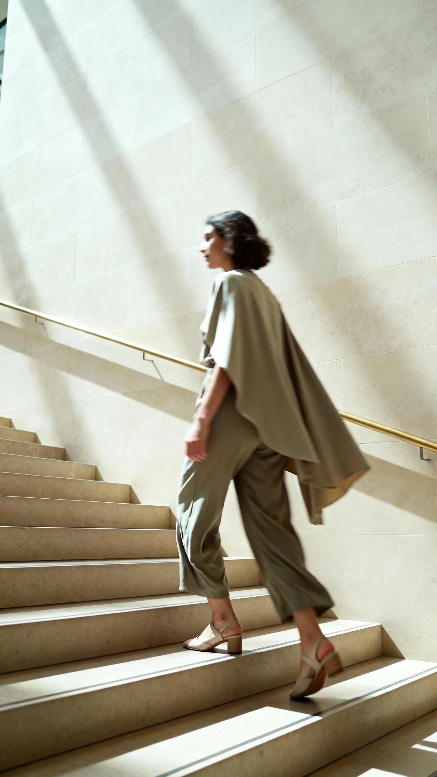 Natural candid of a woman in a flowy jumpsuit with a cape overlay and simple heels, ascending museum steps, face slightly blurred, bright daytime light, iPhone photo quality, unstaged.