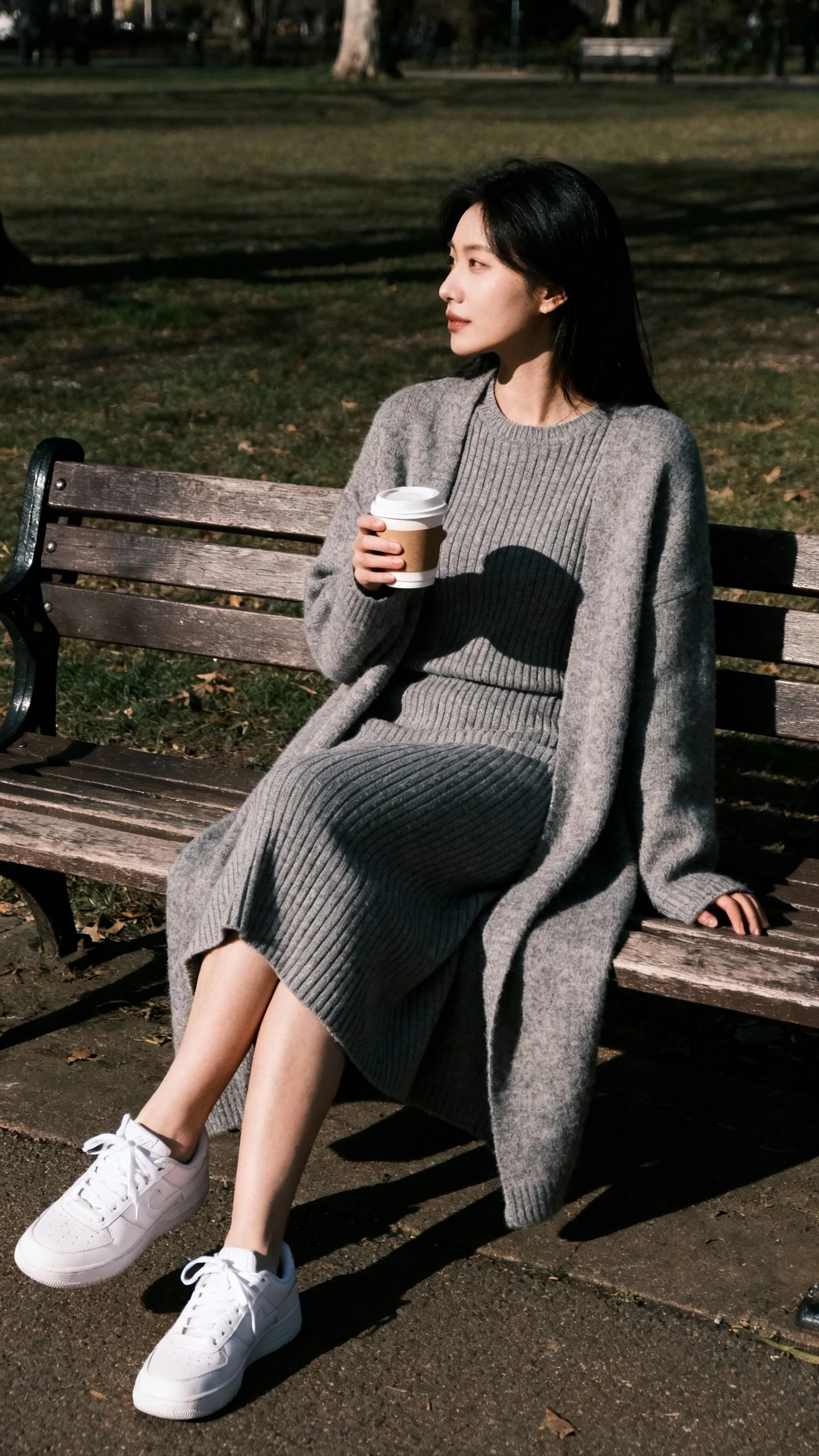 Natural lifestyle photo of a woman wearing a knee-length ribbed knit dress with long sleeves and a cozy fit, layered with a long cardigan and white sneakers, holding a coffee on a park bench, face in shadow looking aside, overcast daylight, iPhone photo quality.