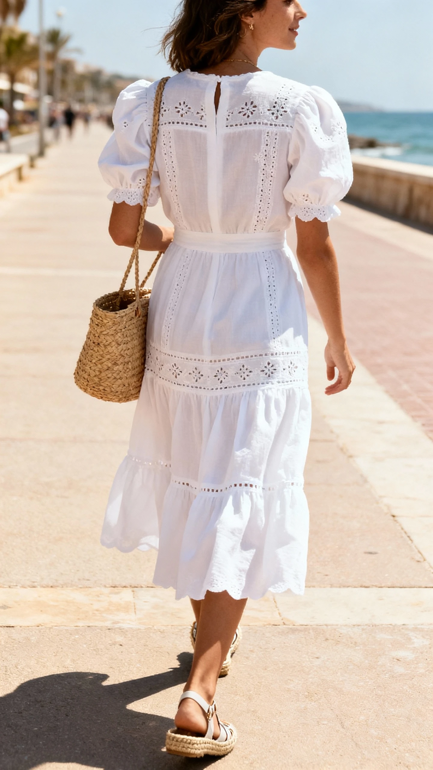 Natural lifestyle photo of a woman wearing a knee-length white broderie anglaise dress with puff sleeves and a tiered skirt, woven straw bag and espadrille wedges, walking along a sunlit seaside promenade, face turned away, bright daylight, iPhone photo quality.