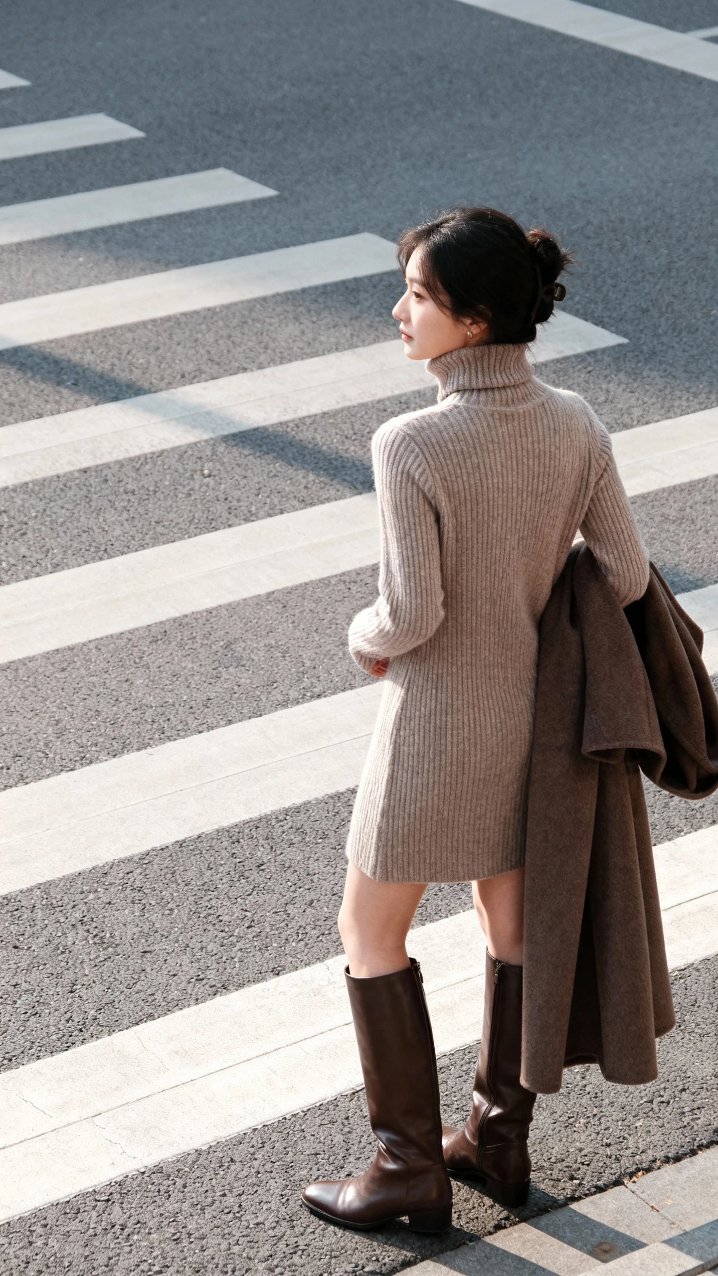 Natural photo of a woman in a ribbed turtleneck sweater dress with knee-high leather boots and a long coat draped over her arm, waiting at a crosswalk, face looking away, soft winter daylight, iPhone snapshot feel.
