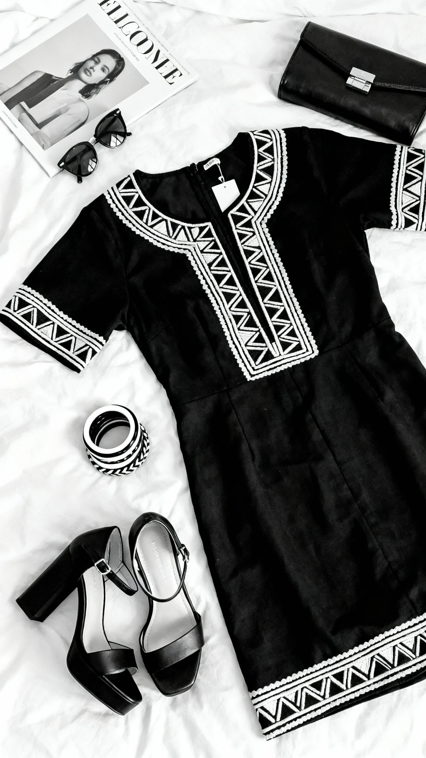 Overhead flat lay of a Xhosa-inspired black-and-white dress with bold geometric trims, block-heel sandals, stack of black-and-white bangles, and a structured black clutch, arranged on a white bedspread with sunglasses and a minimalist magazine, bright natural light, no person visible, organized composition, iPhone photo aesthetic.