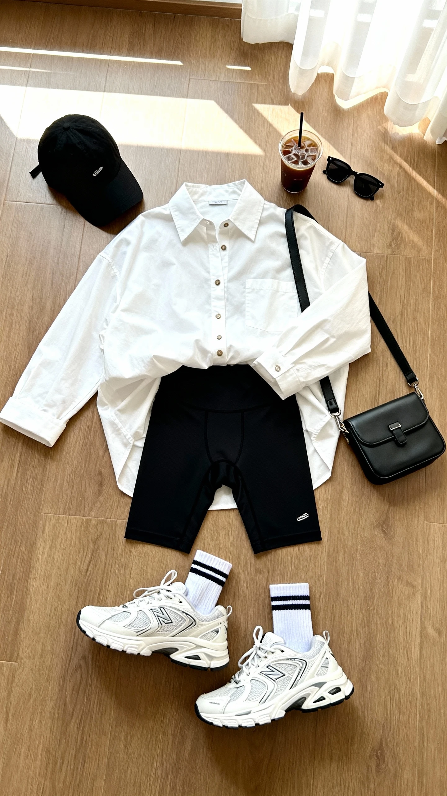 Overhead flat lay of oversized white button-up half-tucked styling with black bike shorts, chunky white trainers, sporty crew socks, black baseball cap, mini crossbody bag, and sleek sunglasses on a wooden floor with an iced coffee as prop, bright natural window light, no person visible, organized composition, iPhone photo aesthetic.