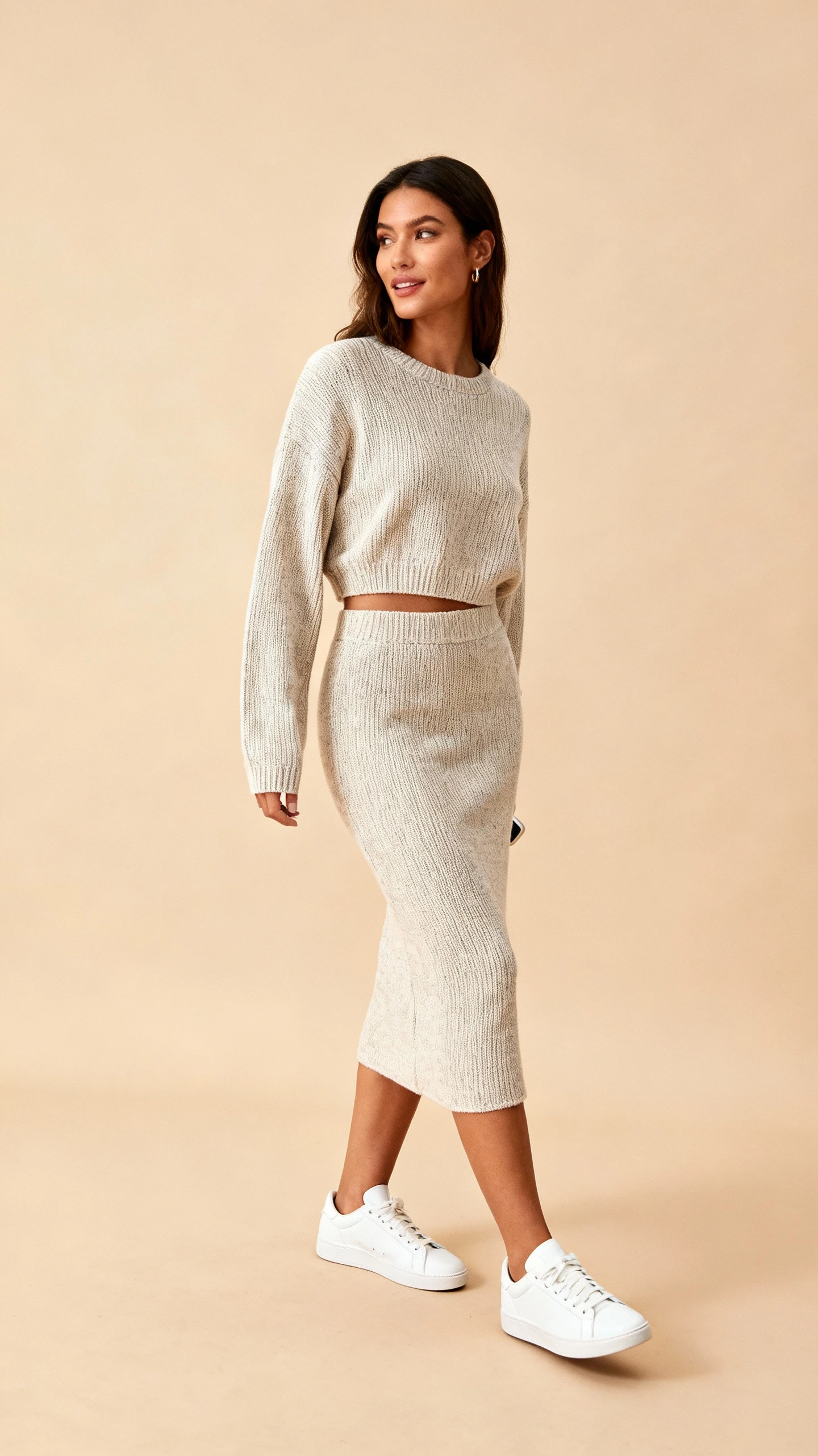 Studio fashion image of a woman in a matching knit top and skirt set styled with classic white sneakers, sand-toned background, soft studio lighting, slight three-quarter turn, clean composition.