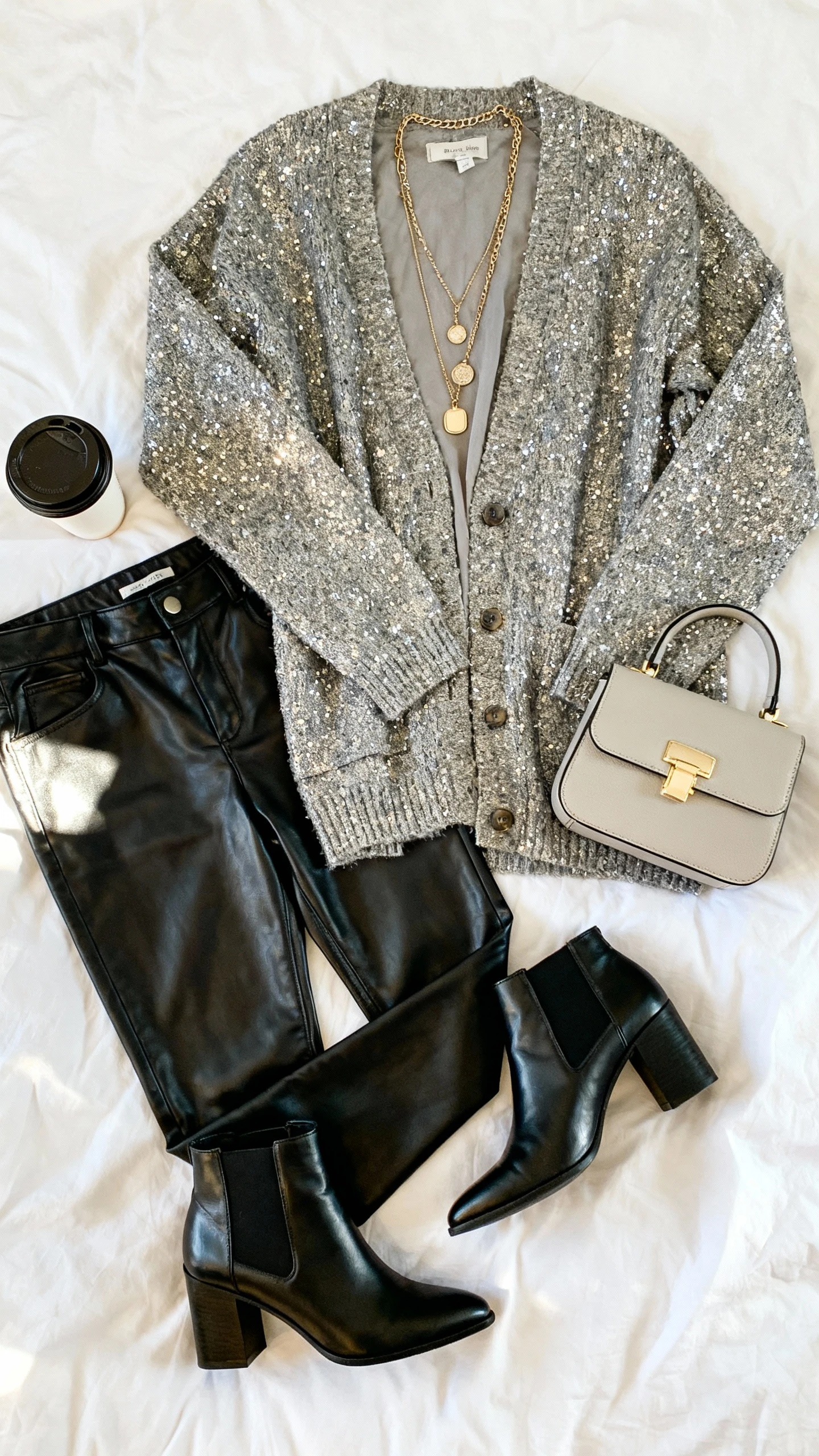 Top-down flat lay of sparkly cardigan, sleek leather pants, ankle boots, layered necklaces, and a mini top-handle bag on white bedspread; add a coffee cup prop, bright natural light, no person visible, organized composition, iPhone photo aesthetic.