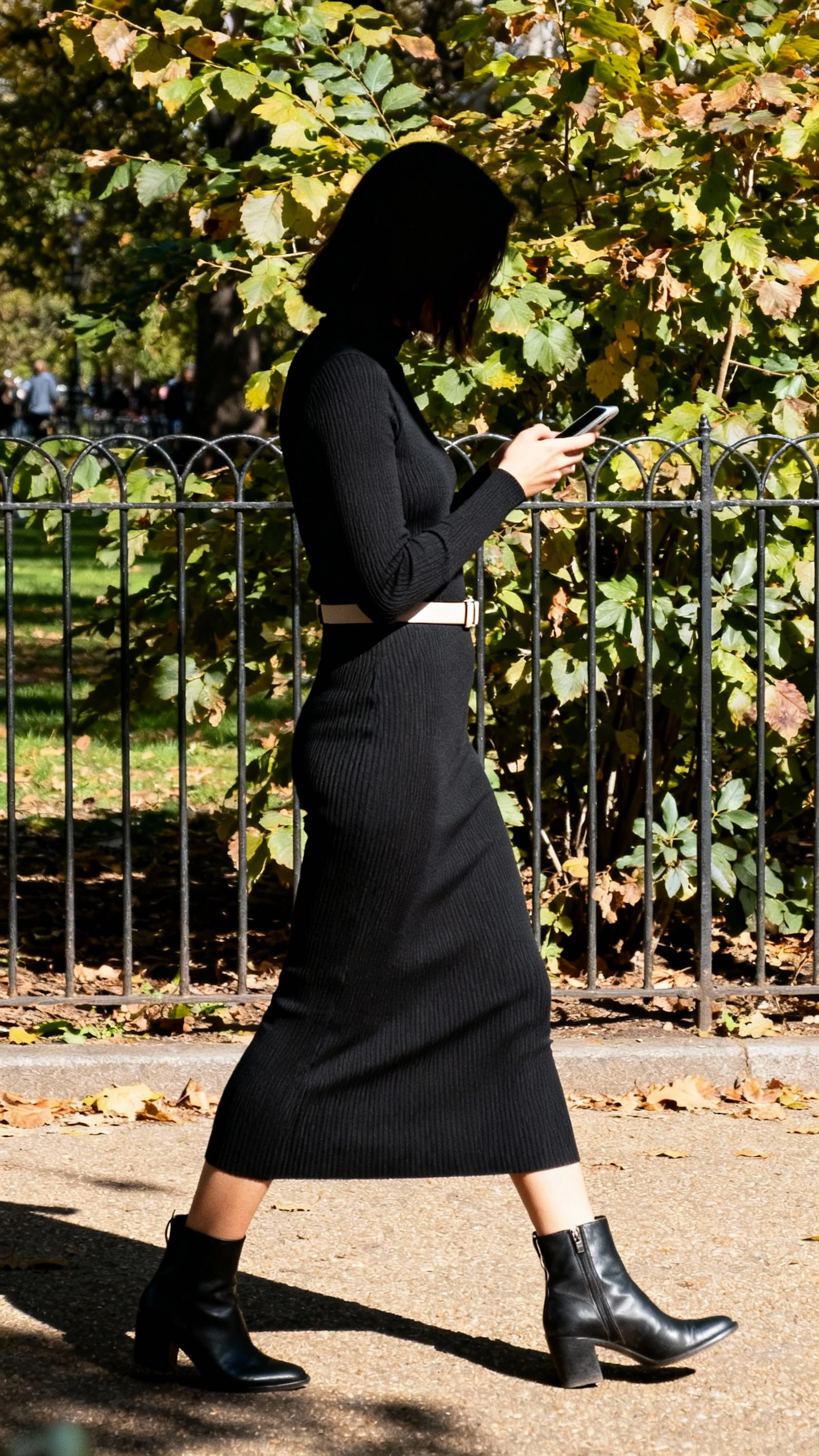 Candid lifestyle shot of a woman in a monochrome knit dress with stretch, minimal belt and ankle boots, walking past a leafy park fence while checking her phone, face in shadow turned away, natural daylight, iPhone photo quality.