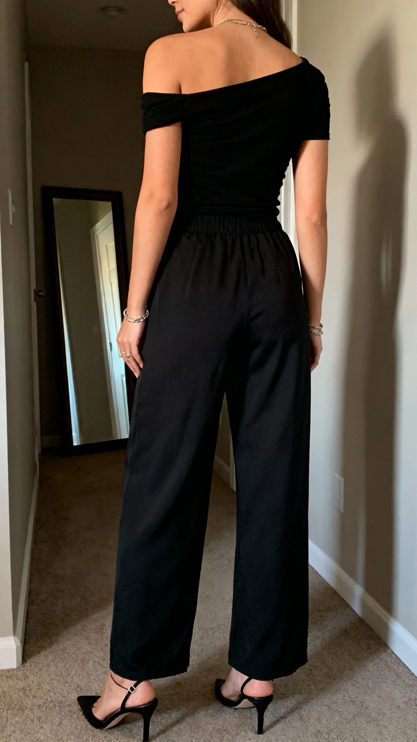 iPhone back view of a woman wearing a black off-shoulder top with fluid high-waisted black trousers, delicate black heels, and understated jewelry, face not visible, hallway mirror, natural evening light, iPhone photo quality.