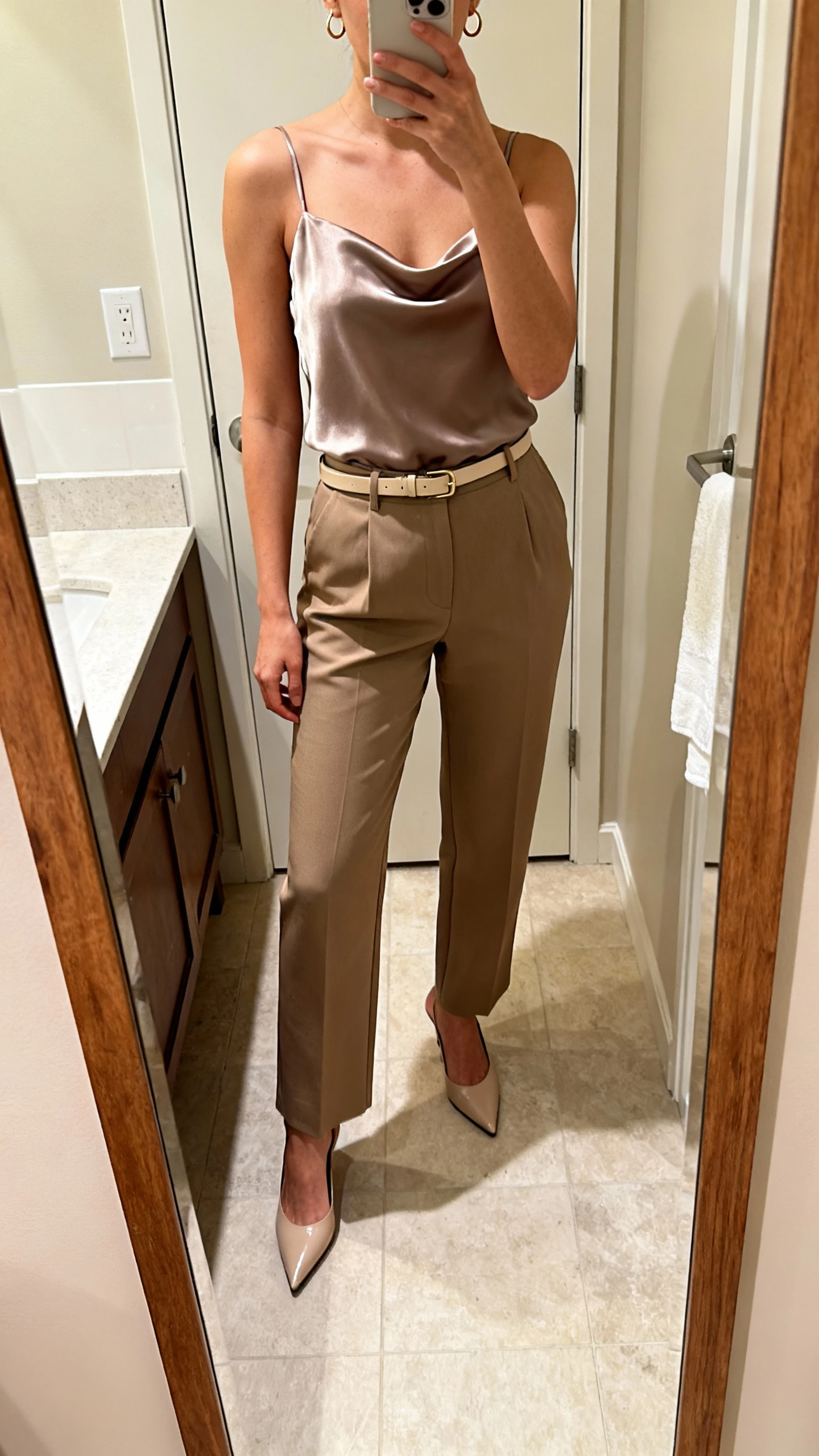 iPhone bathroom mirror selfie of a woman wearing tailored high-waist trousers, a flirty silky camisole top, pointed-toe heels, and a slim belt with small hoop earrings, face not visible, cropped at shoulders, warm indoor lighting, iPhone photo quality.