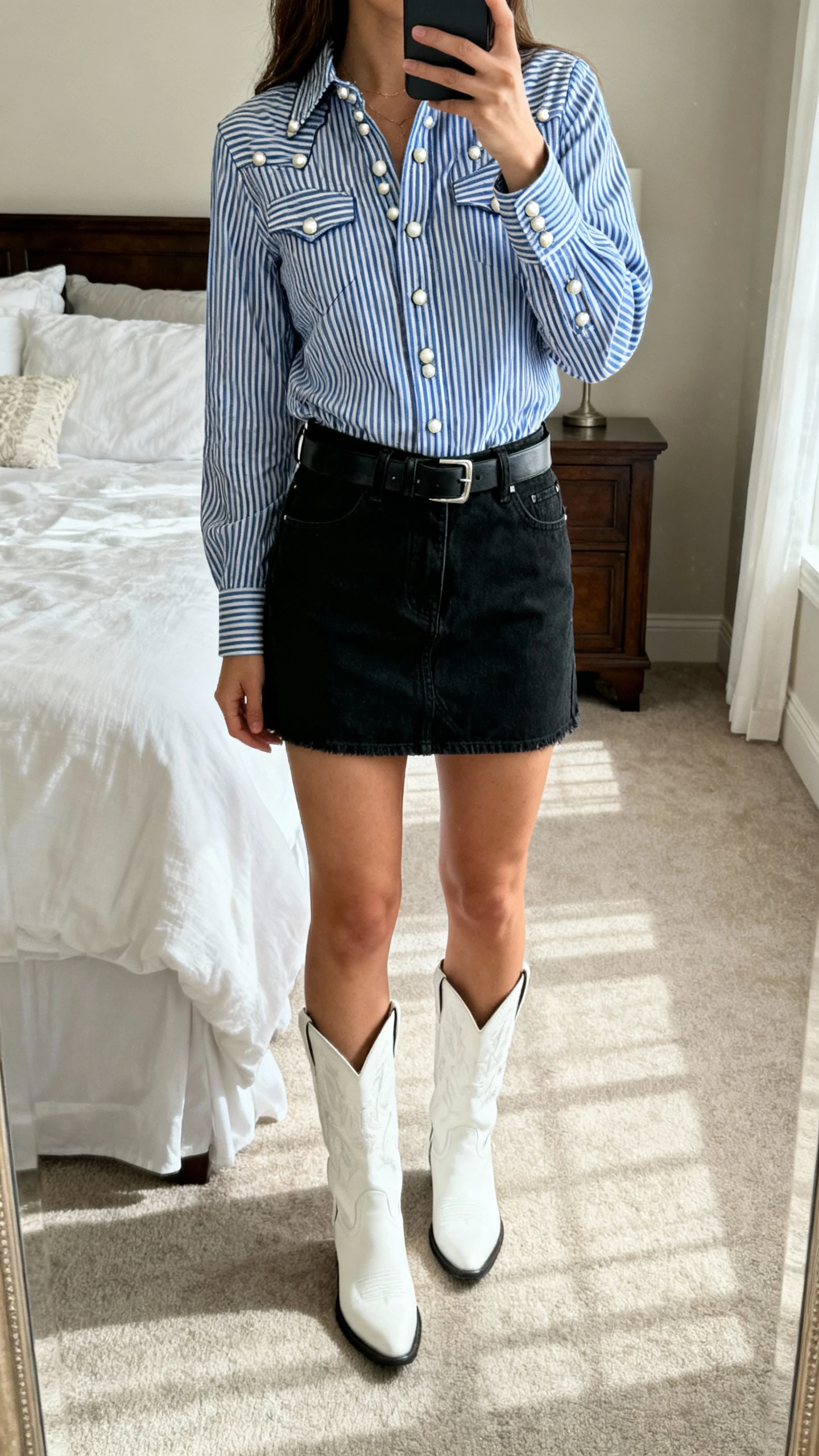 iPhone full-length mirror selfie of a woman in a black denim mini skirt and a blue-and-white striped western button-up with pearl snaps, tucked in, plus white cowboy boots and a black leather belt; face not visible, cropped at shoulders, bedroom mirror, soft daylight, iPhone photo quality.