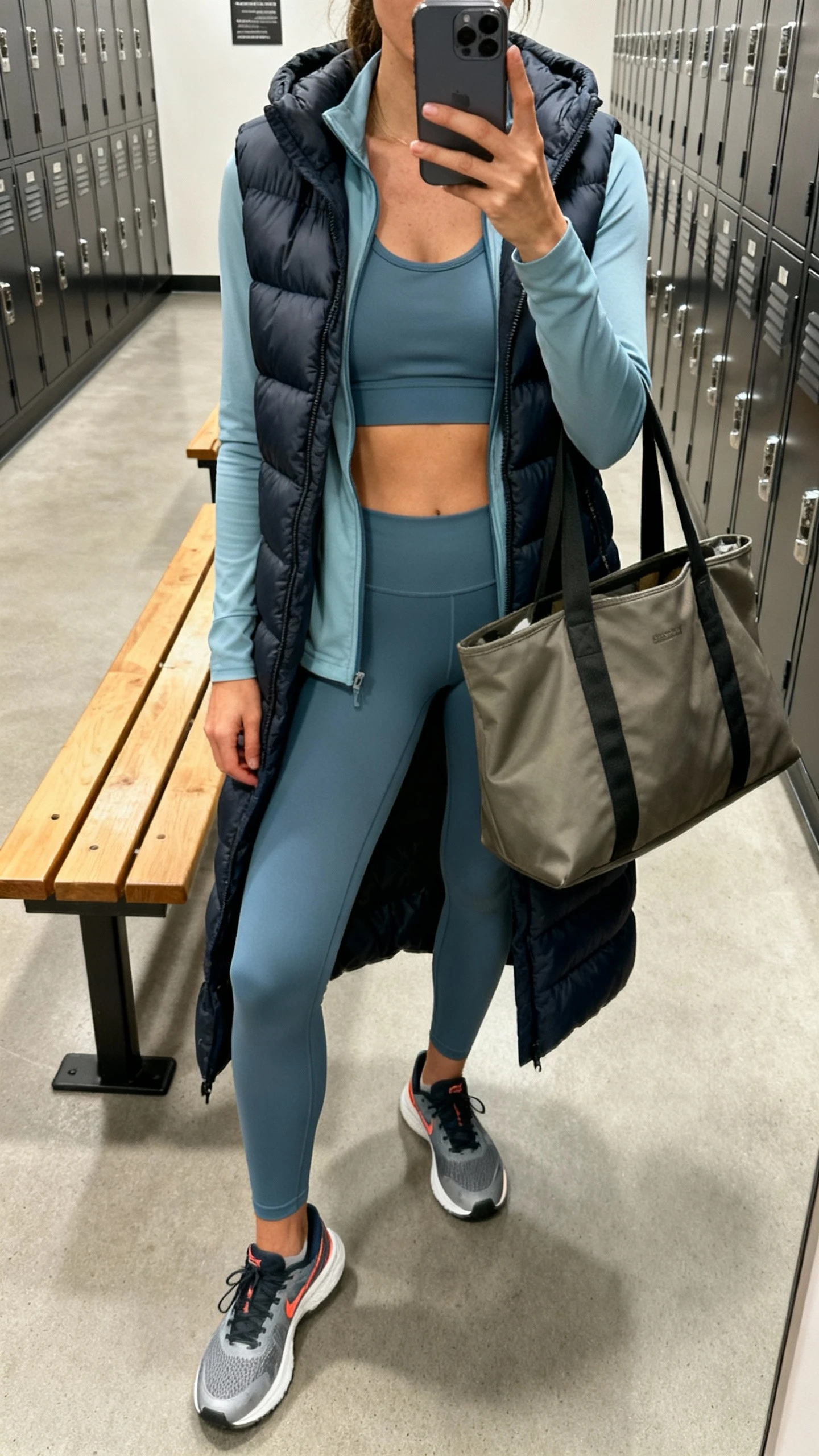 iPhone mirror selfie of a woman in a coordinated athleisure set (sports top and leggings) layered with a long puffer vest, running shoes, large tote, face not visible, gym locker room mirror, natural overhead light, iPhone photo quality.