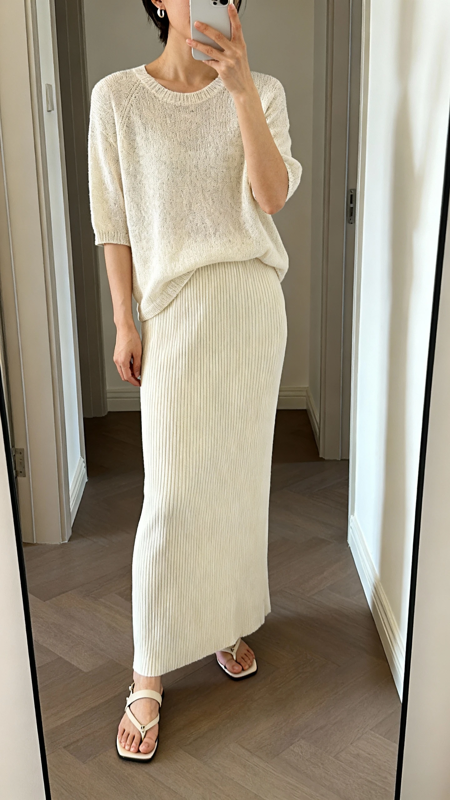 iPhone mirror selfie of a woman in a monochrome look: tonal knit top paired with a matching ribbed long skirt, minimal earrings, and sleek sandals, face not visible, cropped at neck, hallway full-length mirror, soft indoor light, iPhone photo quality.